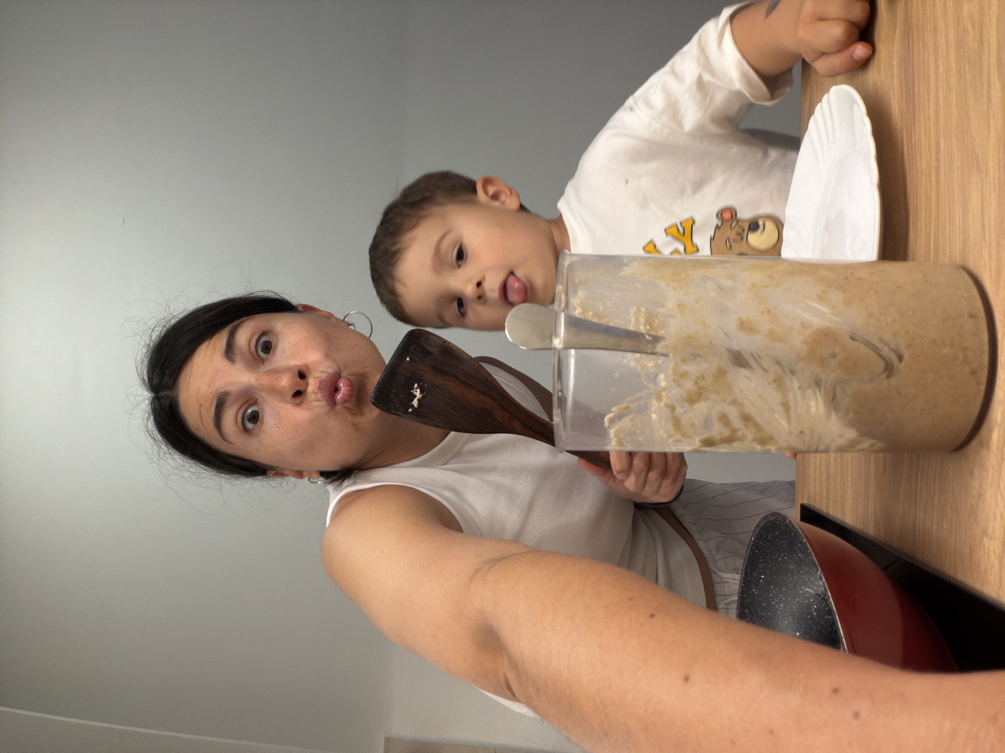 The image captures a playful kitchen scene, featuring a woman and a young child engaged in preparing food. The woman, positioned in the foreground, has dark hair and wears a white sleeveless top. She looks towards the camera with a humorous expression, making a pouty face with wide eyes. She holds a wooden spatula, which has some light-colored batter on its end, and is also holding a large clear container filled with a similar thick mixture, likely batter or dough, with a metal spoon inside it.
Behind and slightly to the right of the woman, a young child with light brown hair peers over a wooden countertop. The child wears a white long-sleeved shirt adorned with a brown teddy bear graphic and partially visible yellow text that reads "LY". His tongue is slightly out, conveying a curious or playful expression as he looks towards the camera.
The activity appears to be a family cooking or baking session. The setting is a kitchen or food preparation area, indicated by the countertop, cooking utensils, and ingredients. A white paper plate stands upright behind the clear container, and a red frying pan with a black speckled interior is visible in the lower right portion of the frame. The background is a plain light gray wall. The overall atmosphere is candid and lighthearted, suggesting a joyful moment between the woman and child in Budva, Montenegro.