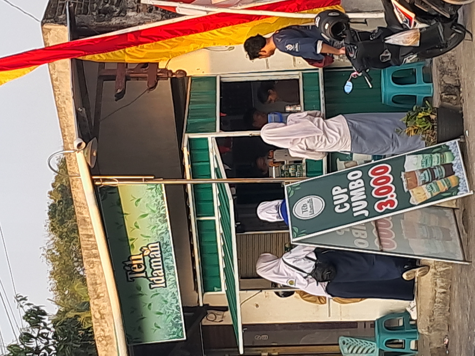A street-level, medium shot captures a small, outdoor food stall with a green awning and a sign advertising "Teh Idaman" and "CUP JUMBO 3.000" in Indonesian. The stall is situated in the city of Ponjong, Indonesia. Several individuals are present. Two young women wearing hijabs and school uniforms stand in front of the stall, facing away from the camera. One has a black bag slung over her shoulder. A man in a blue shirt is standing to the right of the stall, looking towards it. Inside the stall, through an open window, two people appear to be working or serving customers. A parked motorcycle is visible on the right side of the frame. A small plant in a black pot sits near the motorcycle. The background shows a building and foliage, with a flag featuring red and yellow stripes to the right of the stall. The overall scene depicts a typical street-side business in Indonesia.