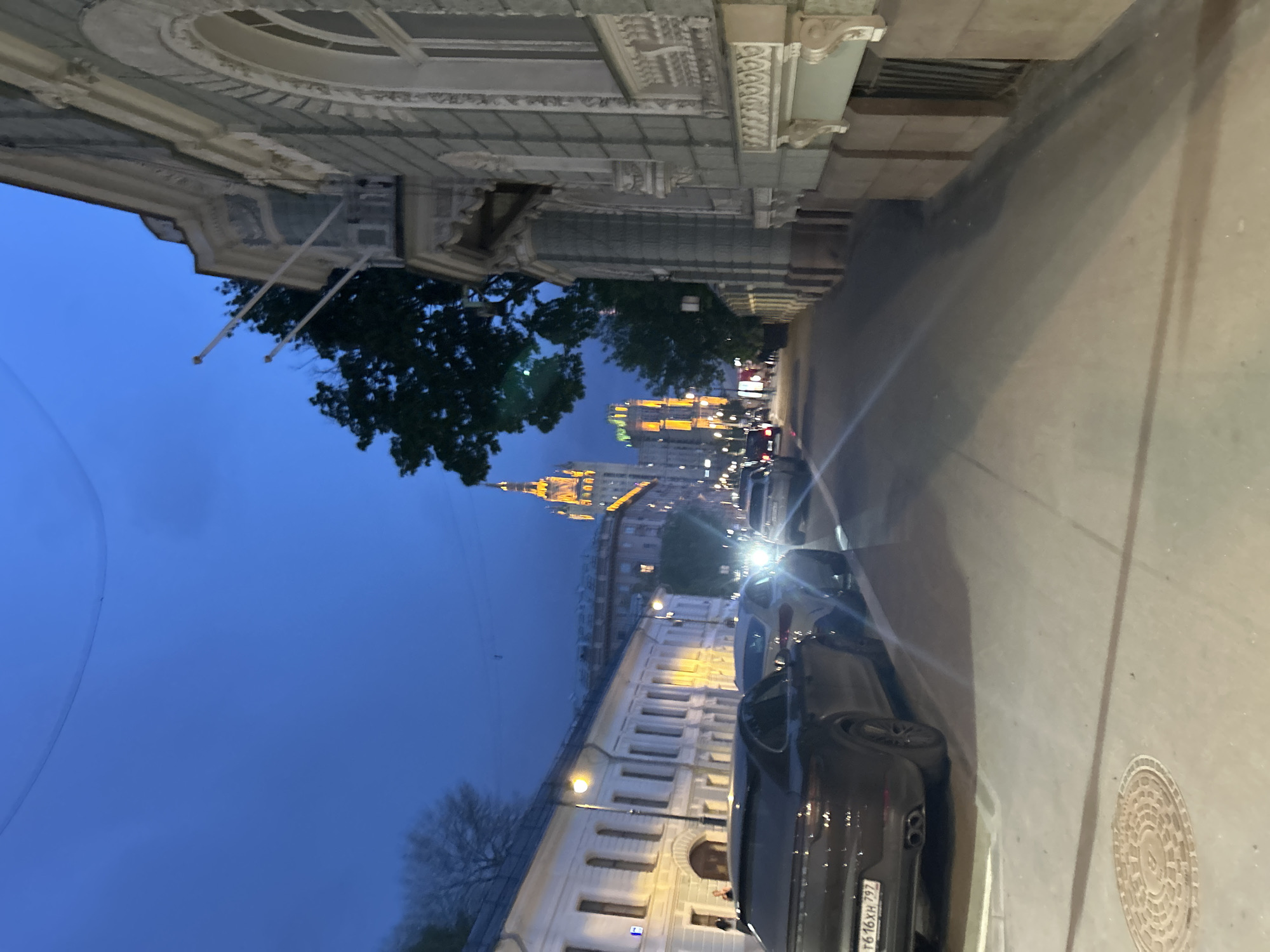 Captured from ground level in Unknown, Unknown, the image presents a street view at dusk. A dark-colored car is parked on the right side of the street, its license plate visible. A manhole cover is located in the street near the car. Buildings line both sides of the street, featuring architectural details and street lighting. In the distance, a brightly illuminated building, possibly a skyscraper or university, rises above the other structures. The sky transitions from light to dark blue, indicating a time shortly after sunset. Trees are visible between buildings, adding a touch of nature to the urban landscape.