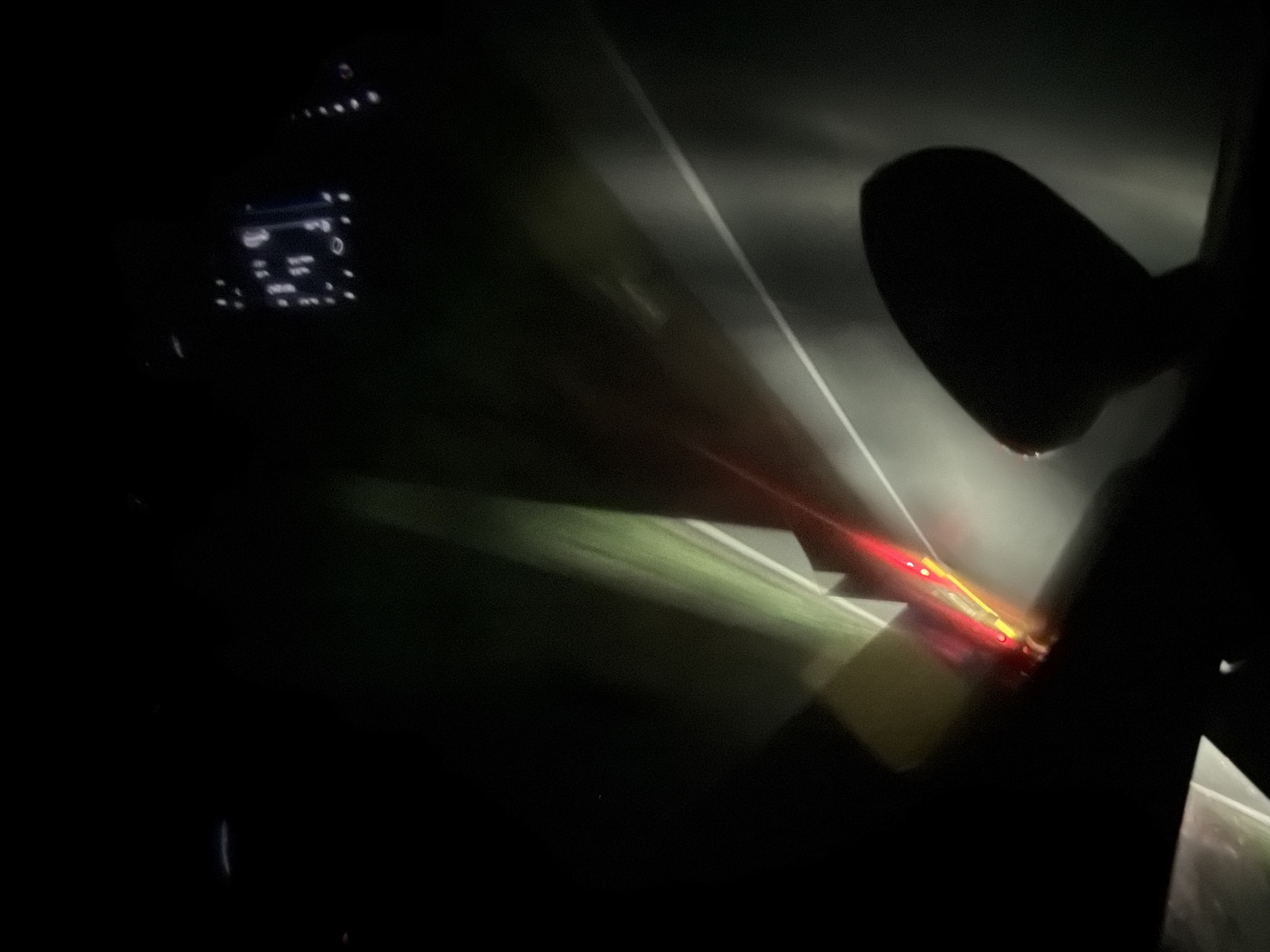 The image depicts a night-time view from inside a vehicle, presumably in Trudovets, Bulgaria. The scene is dark, with the primary light sources coming from the vehicle's interior, dashboard lights, and exterior lights from other vehicles or roadside illumination. In the foreground, a dark silhouette of a rearview mirror is visible. The road ahead is dimly lit, with a white line marking the lane. A red and yellow light source, likely from another vehicle's taillights or a roadside signal, is visible in the middle distance, creating a streak of light. The overall impression is one of driving at night through an urban or suburban area.