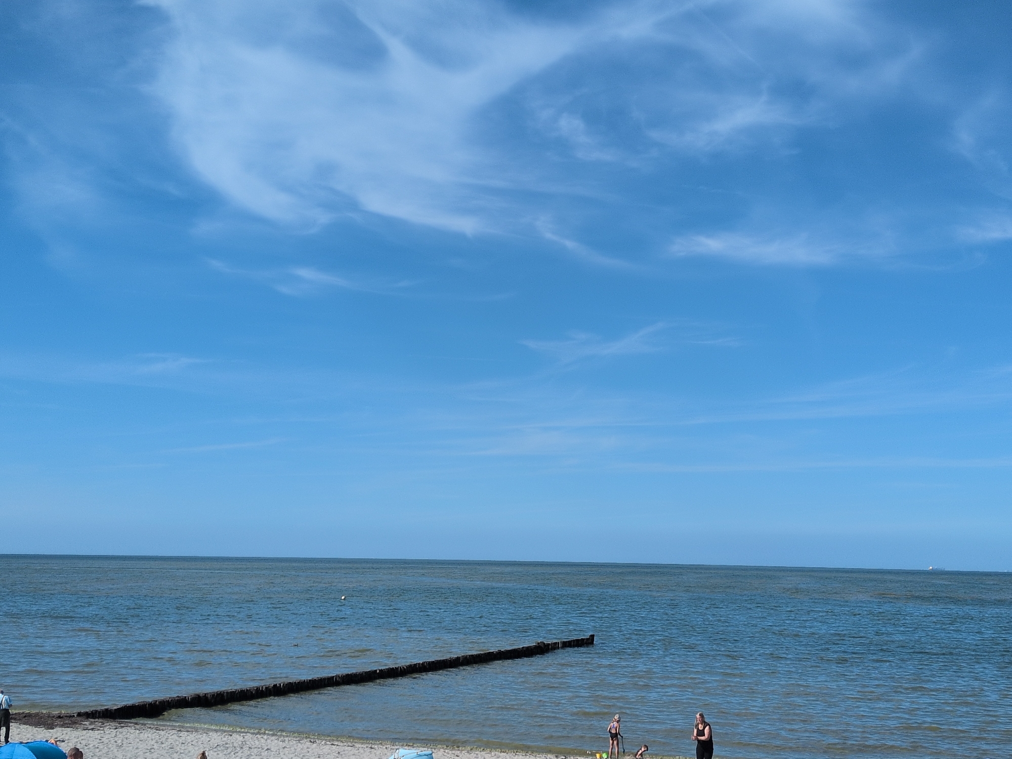 This image depicts a beach scene in Kühlungsborn, Germany, on a clear, sunny day with a blue sky filled with wispy white clouds. The foreground features a sandy beach with several people. Two women are visible: one in a black tank top and shorts, standing near the water's edge, and another in a black swimsuit, also near the water. A child is visible in the sand between them, appearing to be playing. Further to the left, partially obscured by a blue object, is a person in dark pants and a lighter shirt, facing away from the camera.
The sea stretches out to the horizon, appearing calm with gentle waves. A long wooden breakwater extends into the water from the left side of the frame, creating a barrier against the waves. A small white buoy floats in the water to the left of the breakwater. In the distance, a ship can be seen on the horizon. The overall impression is a peaceful summer day at the seaside.