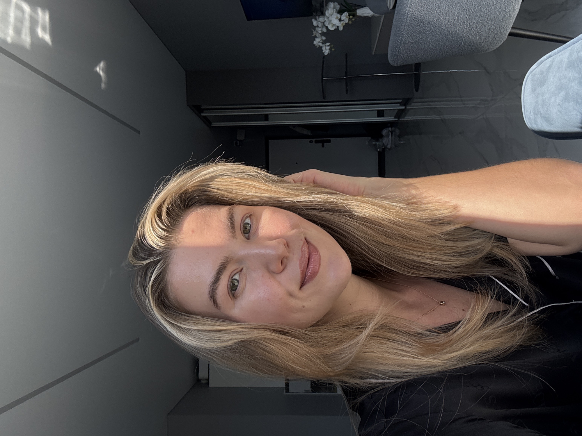A woman with blonde hair is positioned in an indoor setting. She is fair-skinned, with green eyes and a slight smile. She is wearing a dark shirt and a delicate necklace. Her hand is touching her hair. The background includes a gray wall with a subtle linear design, a chair, a decorative arrangement of white flowers, and what appears to be a metallic structure or rack. The location is in an unspecified city, Unknown.