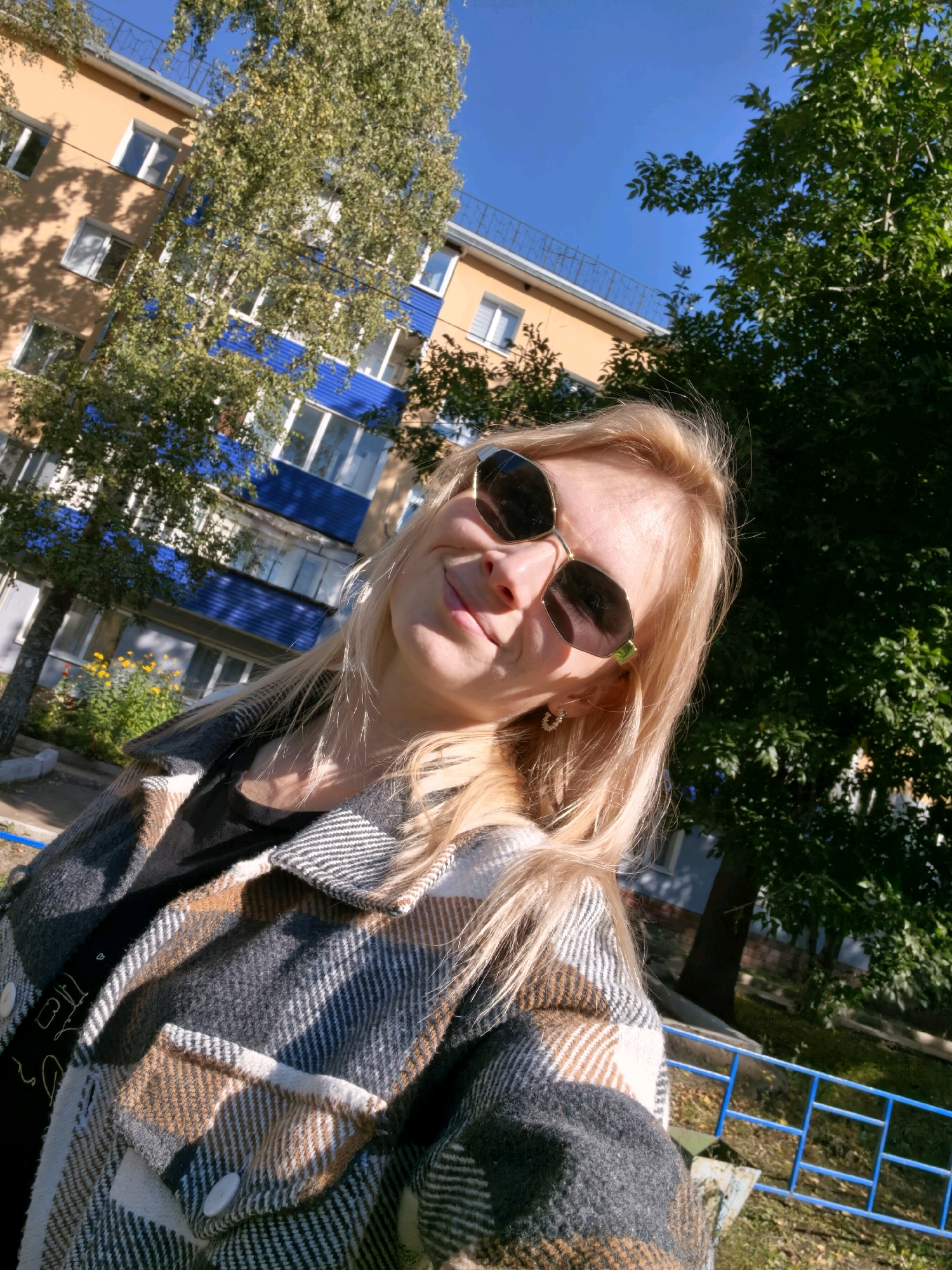 A blonde woman with sunglasses takes a selfie outdoors in front of a building in Sterlitamak, Russia. The woman is wearing a plaid jacket over a dark t-shirt. The building appears to be an apartment complex with orange and blue accents. Trees with green foliage are visible in the background, along with a blue metal fence. The sky is clear and blue. The sun casts shadows on the building and the ground. The image is taken from a low angle, looking up at the woman and the building.