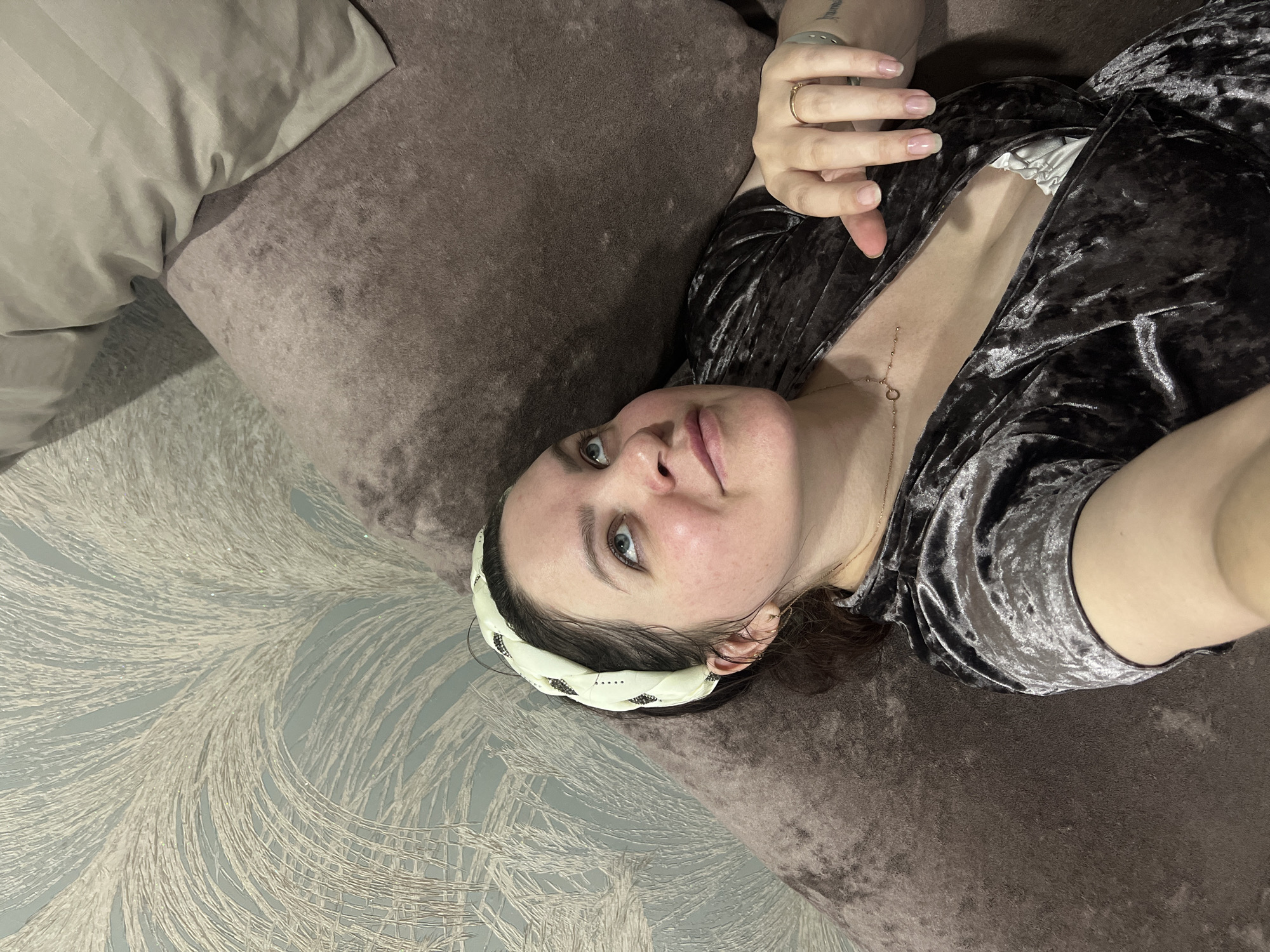 A woman lies on a brown sofa. She wears a dark velvet top and a patterned headband. Her right hand is raised, displaying a ring. The wall is patterned with a white and grey design. The location is not specified in the image, so I cannot confirm if it is Noviy, Russia.