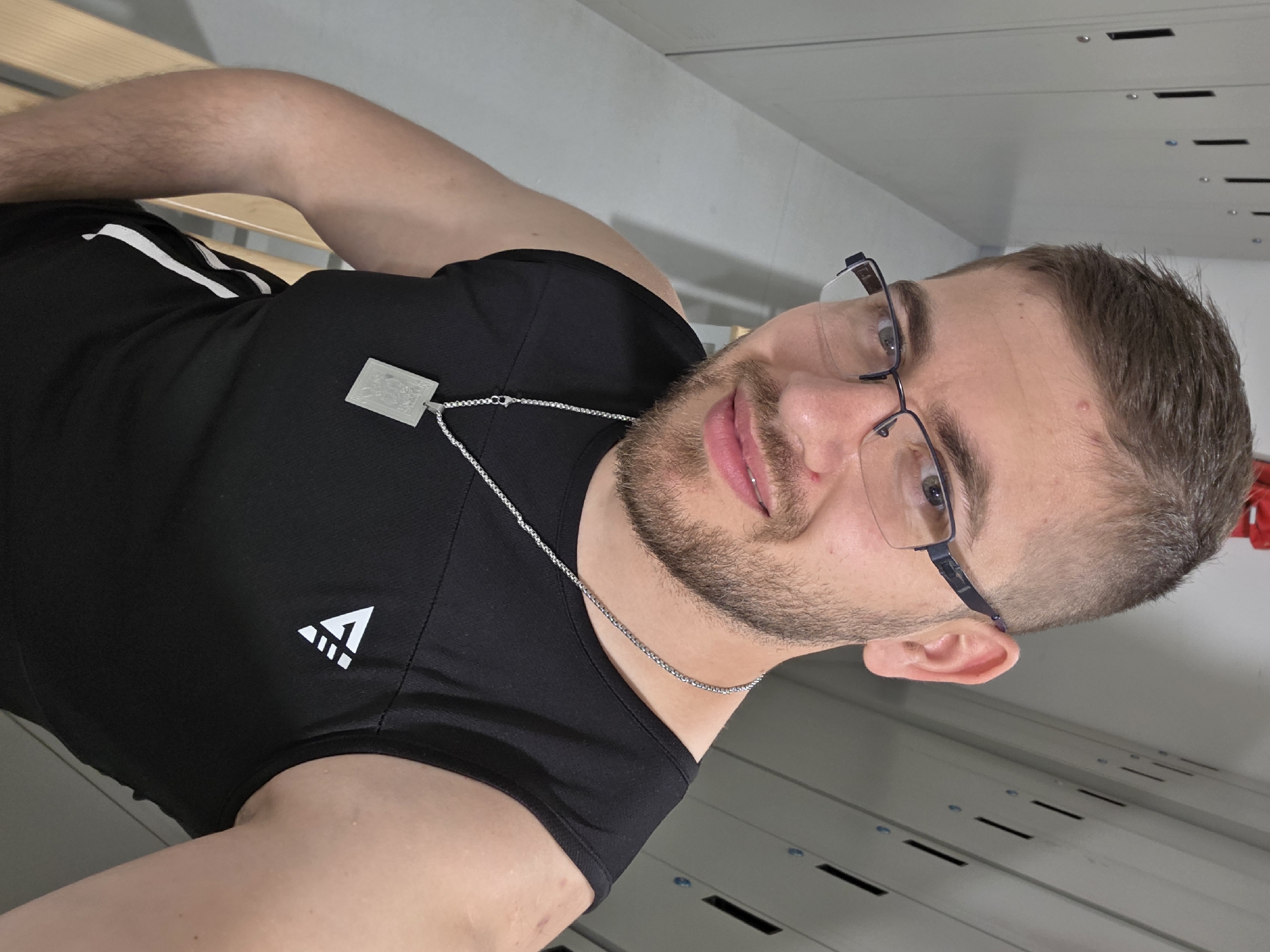A male individual is captured in a selfie, likely taken in an indoor setting such as a locker room or a storage facility in Plovdiv, Bulgaria. The image, originally rotated, shows the person upright. He has short brown hair, a neatly trimmed beard and stubble, and is wearing black-framed glasses. His expression is a slight, friendly smile as he looks directly at the camera.
He is dressed in a black athletic t-shirt and black shorts with white stripes, partially visible on his leg. On the left side of his t-shirt, a white, stylized triangular logo resembling a mountain or the letter 'A' with horizontal lines is visible. Around his neck, he wears a silver chain necklace with a distinctive rectangular, possibly translucent or frosted, pendant. The pendant appears to have an embossed or etched design, though specific details are not clearly discernible.
The background consists of light grey, possibly metallic, panels stacked horizontally, resembling lockers or storage units. These panels feature small dark rectangular vents or slots and small bolt-like fasteners, indicating an industrial or functional space. The lighting is uniform, suggesting indoor artificial light. A bare arm and knee are visible in the foreground, indicating a relaxed posture while taking the picture.