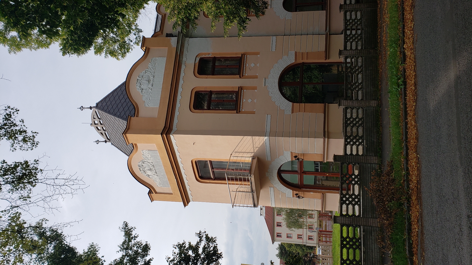 The photograph depicts the facade of a historical building located in the city of Prešov, Slovakia. The building is predominantly peach-colored with white decorative trim. It features an arched entrance at ground level and an arched window above. The building has a distinctive conical roof with decorative elements. A modern-looking balcony with a metal railing extends from the second story. A patterned concrete fence with brick posts separates the property from the street. The street is paved and bordered by a strip of grass and fallen autumn leaves. In the background, another multi-story building with red accents is visible. Trees frame the scene on either side. The sky is overcast.