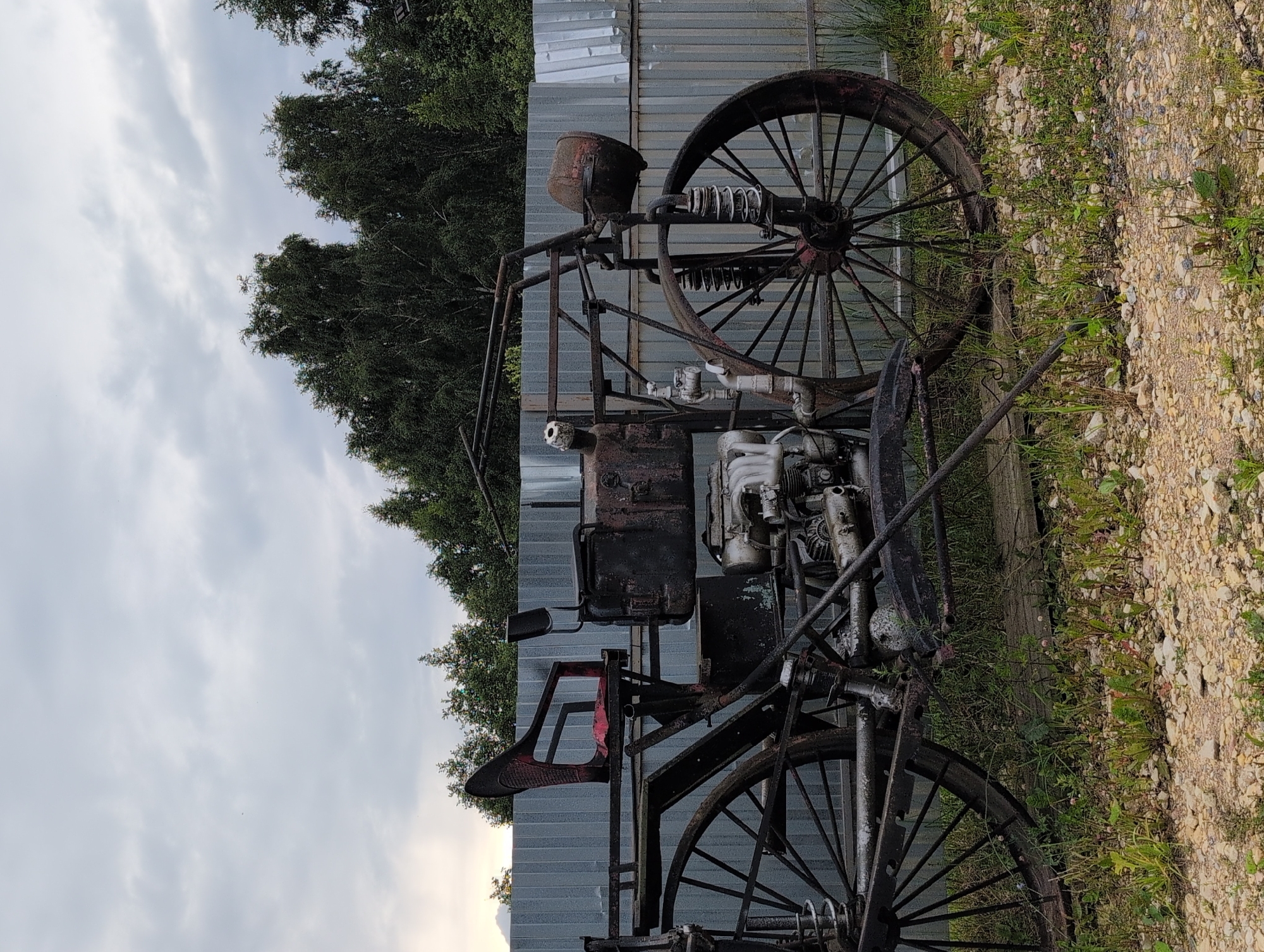 The image depicts an old, rusted, three-wheeled vehicle, possibly a custom-built tricycle or a unique motorcycle. The vehicle is situated outdoors, in what appears to be a rural or semi-abandoned area. It has two large spoked wheels at the rear and one at the front, with a rudimentary engine mounted centrally. The seating appears to be a worn red seat. The background consists of a corrugated metal fence, lush green trees, and an overcast sky. The ground is covered in gravel and overgrown vegetation. The location is specified as Novoe Devyatkino, Russia. No people are visible in the image.