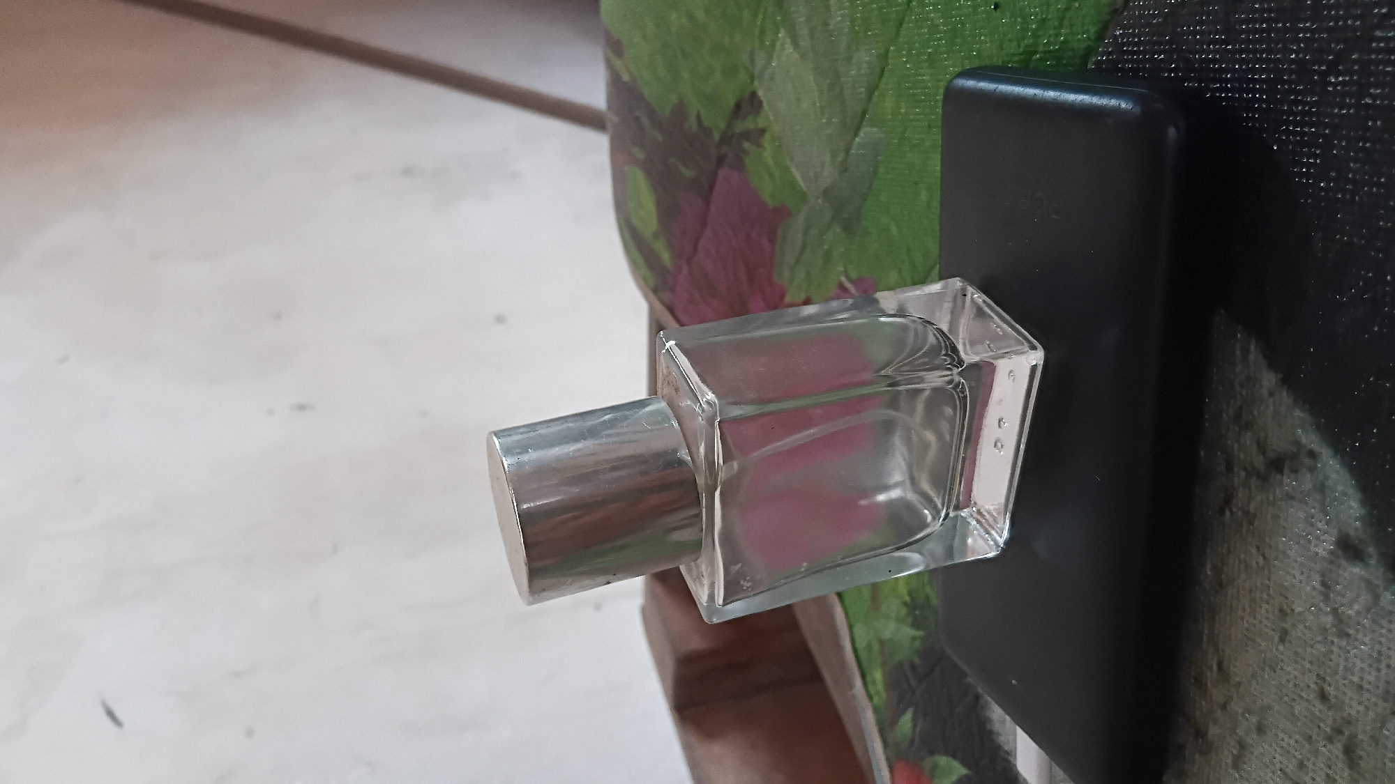 A clear glass perfume bottle with a silver cap is tilted on its side, resting on a black rectangular object. The perfume bottle is square-shaped and has a pinkish hue inside. The black object, possibly a power bank or a phone, is placed on a surface with a textured, dark pattern. Behind the perfume bottle, a green object with purple floral patterns is visible, partially obscured by a brown, textured surface. The background is a lightly textured, off-white wall. The scene appears to be indoors, likely in a domestic setting within the city of Prabumulih, Indonesia.