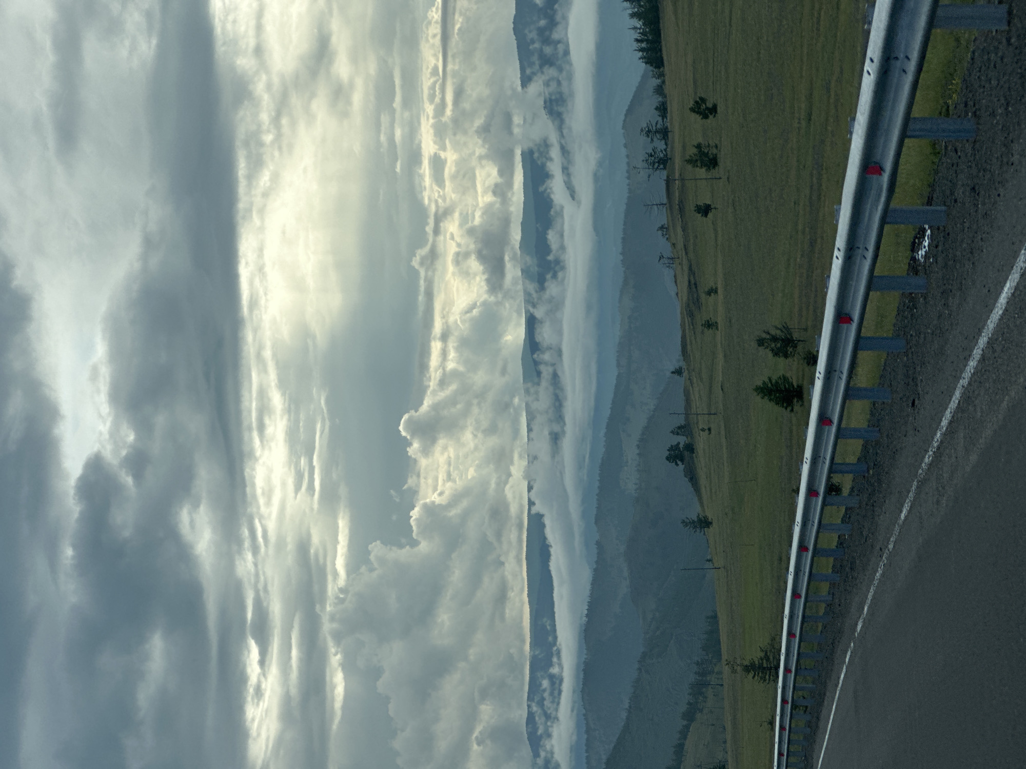 The image shows a view of rolling hills covered in sparse vegetation and scattered trees. A guardrail runs along the side of a paved road in the foreground. In the distance, layers of mountains are visible, partially obscured by clouds and mist. The sky is overcast with dramatic cloud formations. The scene appears to be located in the vicinity of Sakhiurta, Russia, a settlement on Olkhon Island in Lake Baikal. The time of day appears to be late afternoon or early evening, given the soft, diffused light. There are no people or animals visible in the image. The overall atmosphere is one of natural beauty and solitude.