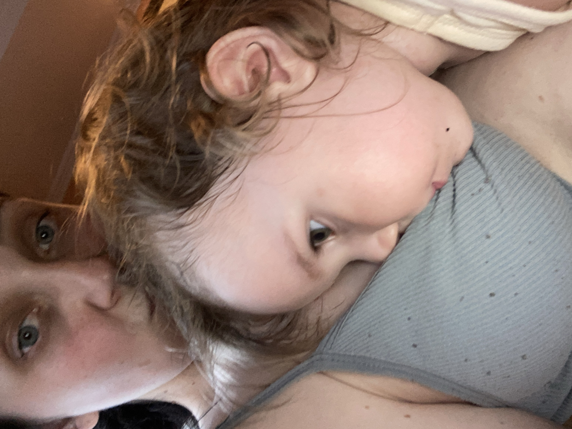 This is a close-up, eye-level, medium shot of a parent and a baby. The parent's face is on the left side of the frame, with their eyes looking towards the camera. The baby is nestled against the parent's chest, with their head turned away from the camera. The baby's hair is a light brown, and their skin is fair. The parent's hair is dark and appears to be long.
The baby is wearing a light-colored shirt, and the parent is wearing a gray tank top. The background is out of focus, but it appears to be a warm, indoor setting. The lighting is soft and diffused, suggesting it might be daytime. There is a sense of intimacy and comfort in the image, with the baby appearing to be asleep or resting peacefully. The parent's expression is gentle and loving.
There are no visible text elements in the image. The overall mood is one of tenderness and connection.