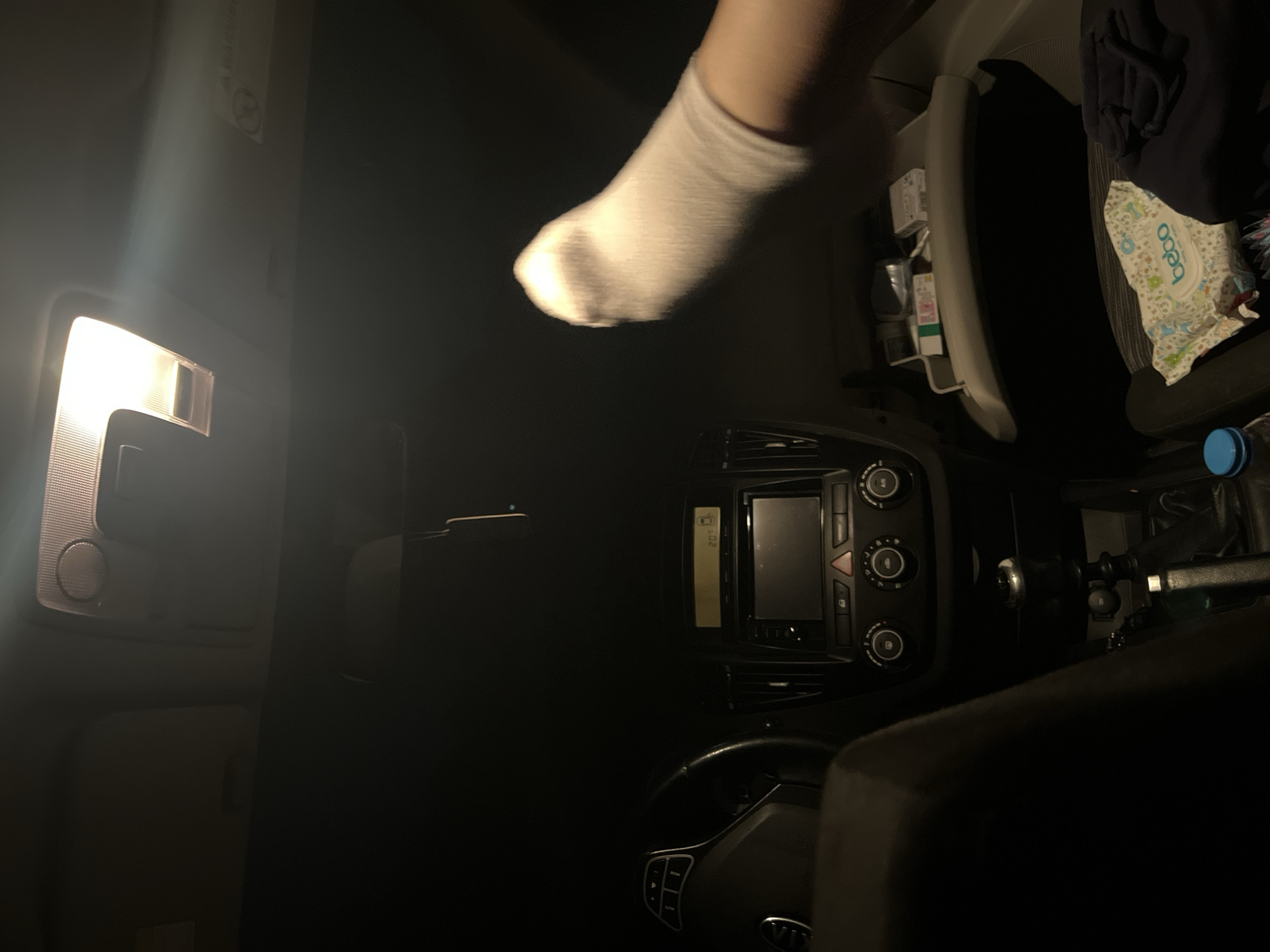 The interior of a Kia car is depicted in low lighting conditions. A foot, covered by a white sock, is visible near the glove compartment. The dashboard, steering wheel, and center console with its controls are discernible. Baby wipes are visible in the passenger seat. No specific location in Marulevo, Bulgaria, can be determined from the image.