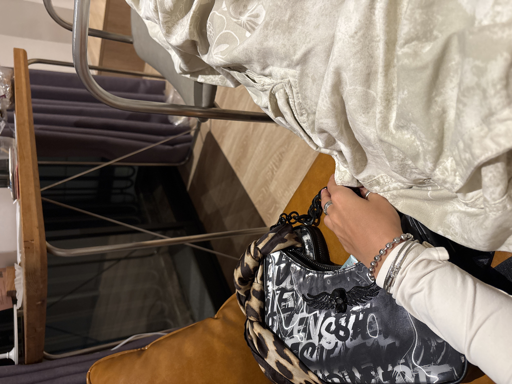 An overhead shot shows a person's arm resting on a bag and draped over a blanket. The arm is adorned with several silver bracelets and rings. The bag is black with graffiti-style lettering and a leopard print scarf draped over it. The person is wearing a white long-sleeved shirt. The background consists of a wooden table and a chair with a metallic frame. The setting appears to be indoors, possibly a cafe or a home.