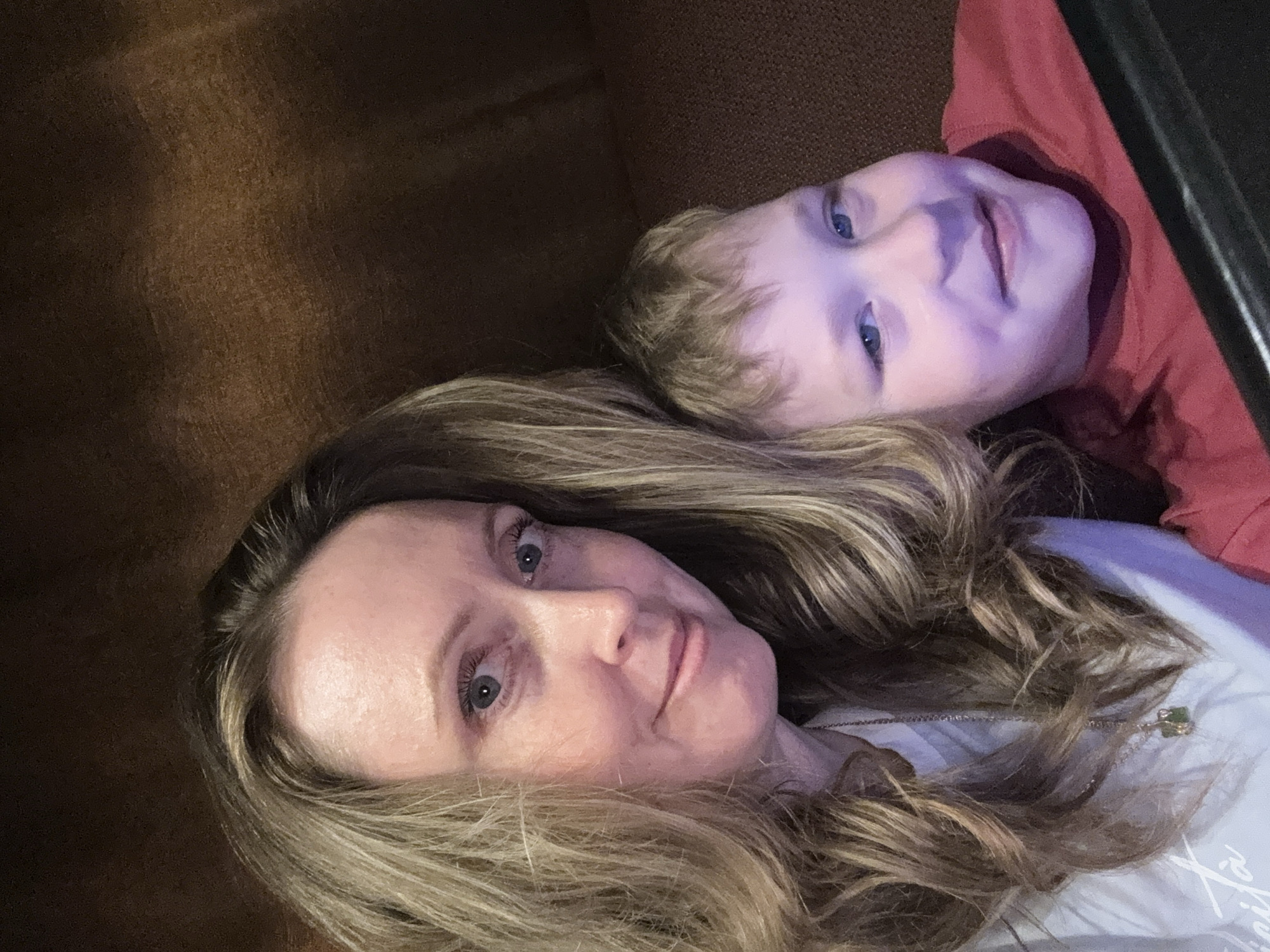 A selfie-style image shows a woman and a young child lying down. The woman has long blonde hair, light skin, and blue eyes. The child also has light skin, blonde hair, and blue eyes and wears a red shirt. The background is a dark brown surface. There is no indication of a location in Balezino, Russia.