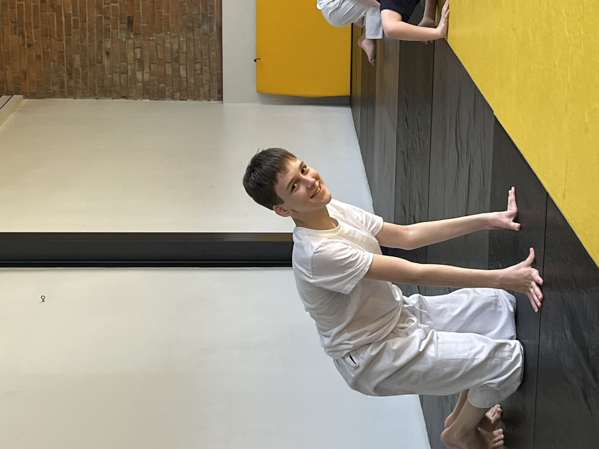 The media file captures an indoor scene, likely a gym or training studio in Krasnoyarsk, Russia. The main subject is a young male, possibly a teenager or young adult, with short dark hair, wearing a white t-shirt and loose-fitting white pants. He is barefoot and smiling broadly at the camera. He is positioned in a low squat or crouch, with both hands pressed flat against a dark wall to his right, suggesting he is either performing a balance exercise, pushing off the wall, or holding a specific pose.
The setting features a mix of architectural elements. The floor appears dark, possibly wood or a textured mat. Directly behind the main subject is a light-colored, possibly white, wall, intersected by a dark horizontal beam or stripe. In the upper left background, a section of exposed red brick wall is visible, hinting at an industrial or older building structure. To the right of the main subject, the wall is predominantly a vibrant yellow, diagonally transitioning into a dark gray or black section at the lower part where his hands are placed. Further in the background, near the top right, another large yellow padded surface or mat is visible.
Parts of other individuals are visible in the background on the right side. These include bare feet and lower legs, also wearing light-colored pants similar to the main subject, and an arm with a hand against the yellow wall. This indicates that other people are present, possibly participating in the same activity or waiting. A small, dark, hook-like object is visible on the light wall in the left background. The overall atmosphere suggests a focused yet enjoyable physical activity session, given the main subject's cheerful expression and the functional setting. There is no visible text in the image.