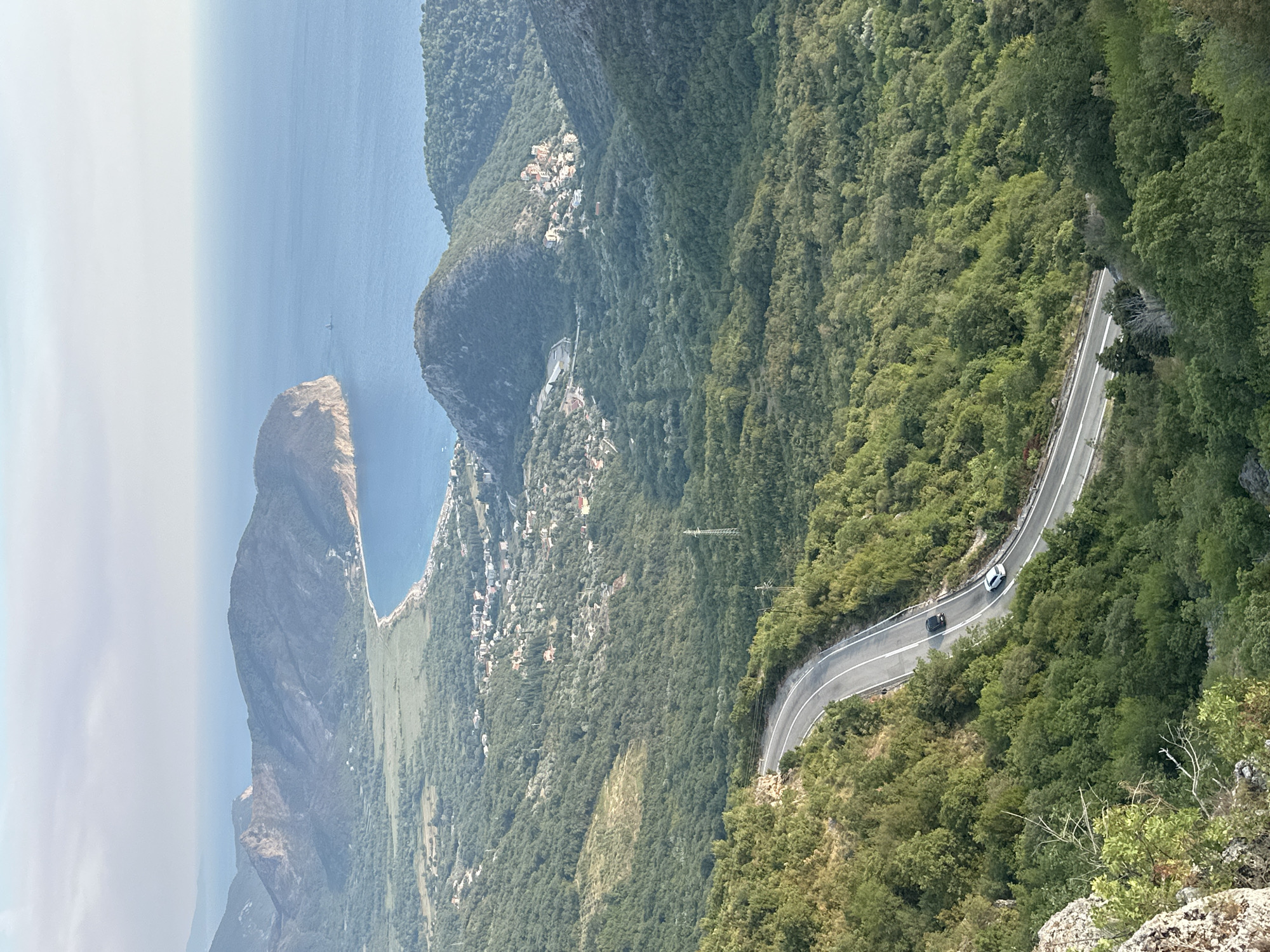 An aerial view captures a winding asphalt road cutting through lush green mountains near Novoselje, Montenegro. Two vehicles, a black car and a white car, are visible on the road. The road curves dramatically as it descends into a valley. In the distance, the Adriatic Sea is visible, meeting a coastline with a small village nestled between the mountains and the water. The sky is hazy, and the overall lighting suggests it is either early morning or late afternoon. The landscape is characterized by dense vegetation covering the rolling hills and mountains.