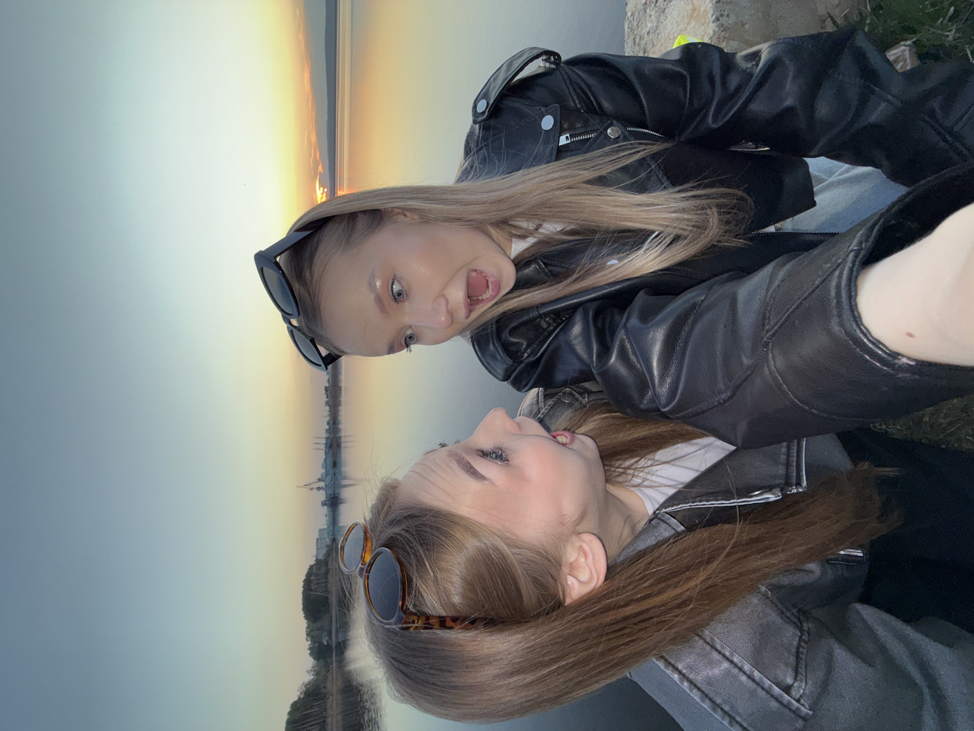 Two young women pose for a selfie against a backdrop of a river or large body of water, likely in Chaikovsky, Russia, suggested by the distant cranes or industrial structures in the background. The blonde woman on the top makes an exaggerated open-mouth expression, while the brunette at the bottom looks up towards her with a slight smile. Both wear sunglasses perched on their heads. They are wearing leather jackets. The scene has a warm glow, suggesting it is either sunrise or sunset. The environment consists of a body of water with an industrial or urban skyline in the background.