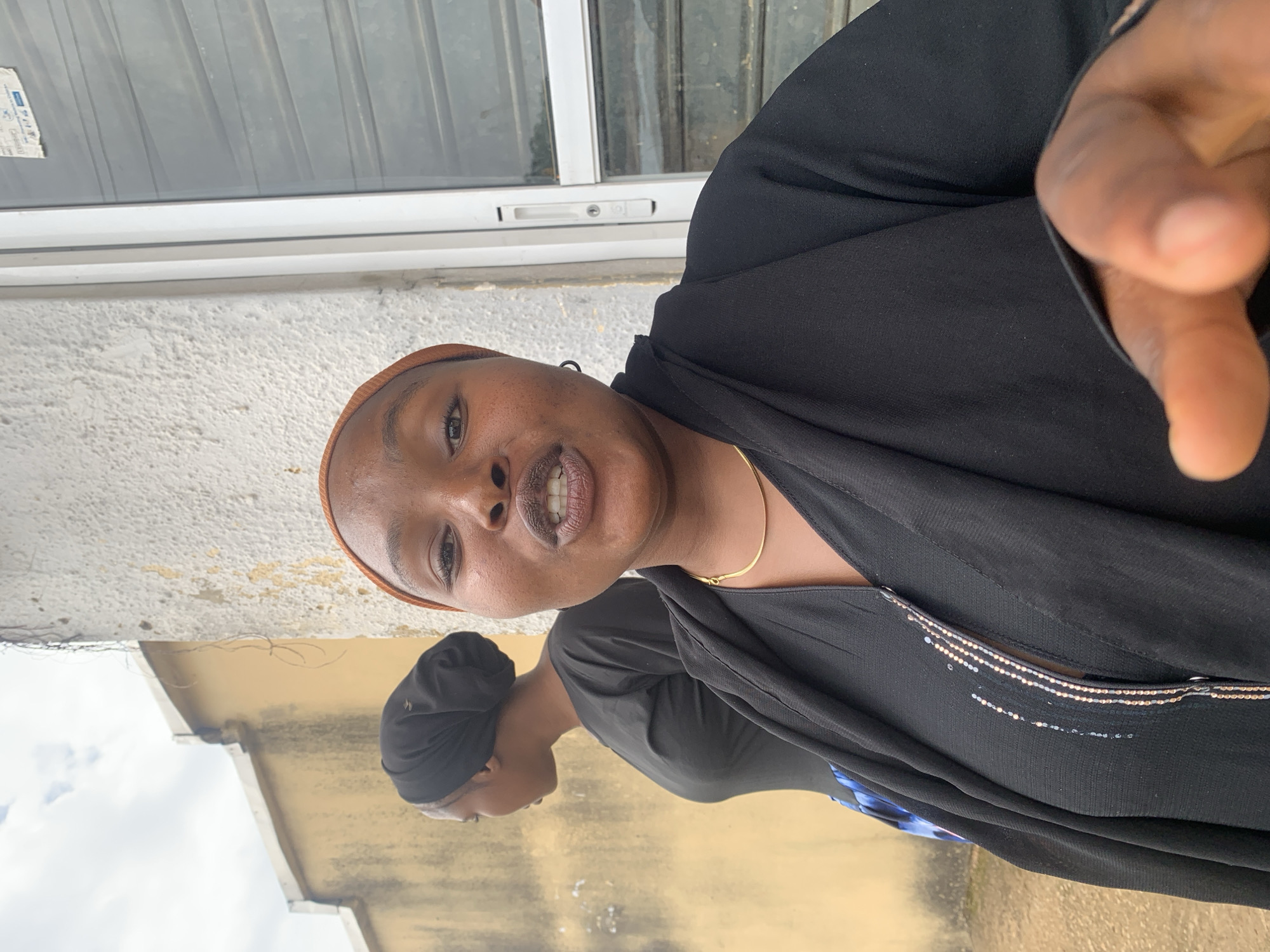 The image depicts an outdoor scene in an urban or semi-urban environment, identified as the city of Jos, Nigeria. In the foreground, a woman with a dark complexion faces the viewer, her mouth slightly open as if speaking, revealing her upper and lower teeth. She wears an orange-brown head covering that frames her face, a thin gold-colored necklace, and a small dark hoop earring on her visible right ear. Her attire consists of a black long-sleeved garment with a central vertical embellishment of shimmering beads or sequins in silver and gold/rose gold tones, covered by an additional loose-fitting black outer garment or shawl. Her right hand is extended towards the camera, with the index finger pointed.
In the background, to the left of the foreground subject, another woman with a dark complexion is seen in profile, looking towards the left side of the frame. She wears a black head wrap and a plain black long-sleeved top, with a segment of a blue and white patterned garment visible below.
The background features two distinct walls: a weathered, light yellow-tan wall on the left behind the second woman, and a textured, off-white concrete wall on the right behind the foreground woman, both showing signs of age and discoloration. Above the yellow wall, an overcast sky is visible. To the far right, a segment of a white window or door frame with a visible handle mechanism and a small label at the top is present, behind which is a dark, possibly shuttered window. Some dry vine-like material hangs from the top of the off-white wall. The ground appears earthy and indistinct at the bottom left.