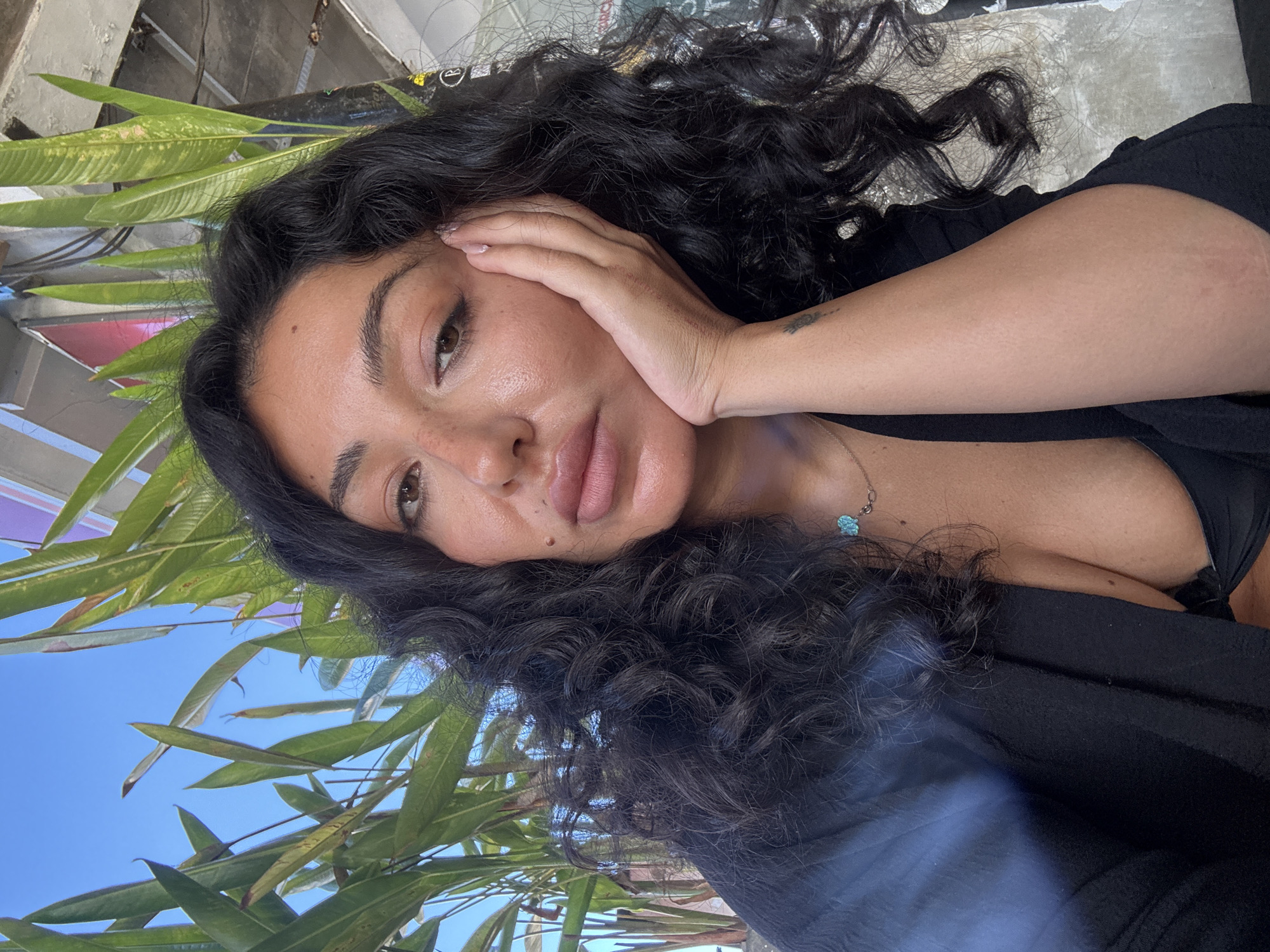 A selfie of a woman with long, dark, curly hair, light brown skin, and brown eyes. She is lying on her side, her head resting on her hand. She is wearing a black top. The background includes green foliage, a blue sky, and indistinct urban elements, suggesting an outdoor location, potentially Kuta Selatan, Indonesia. A silver necklace with turquoise accents sits on her chest. A tattoo is visible on her left forearm.