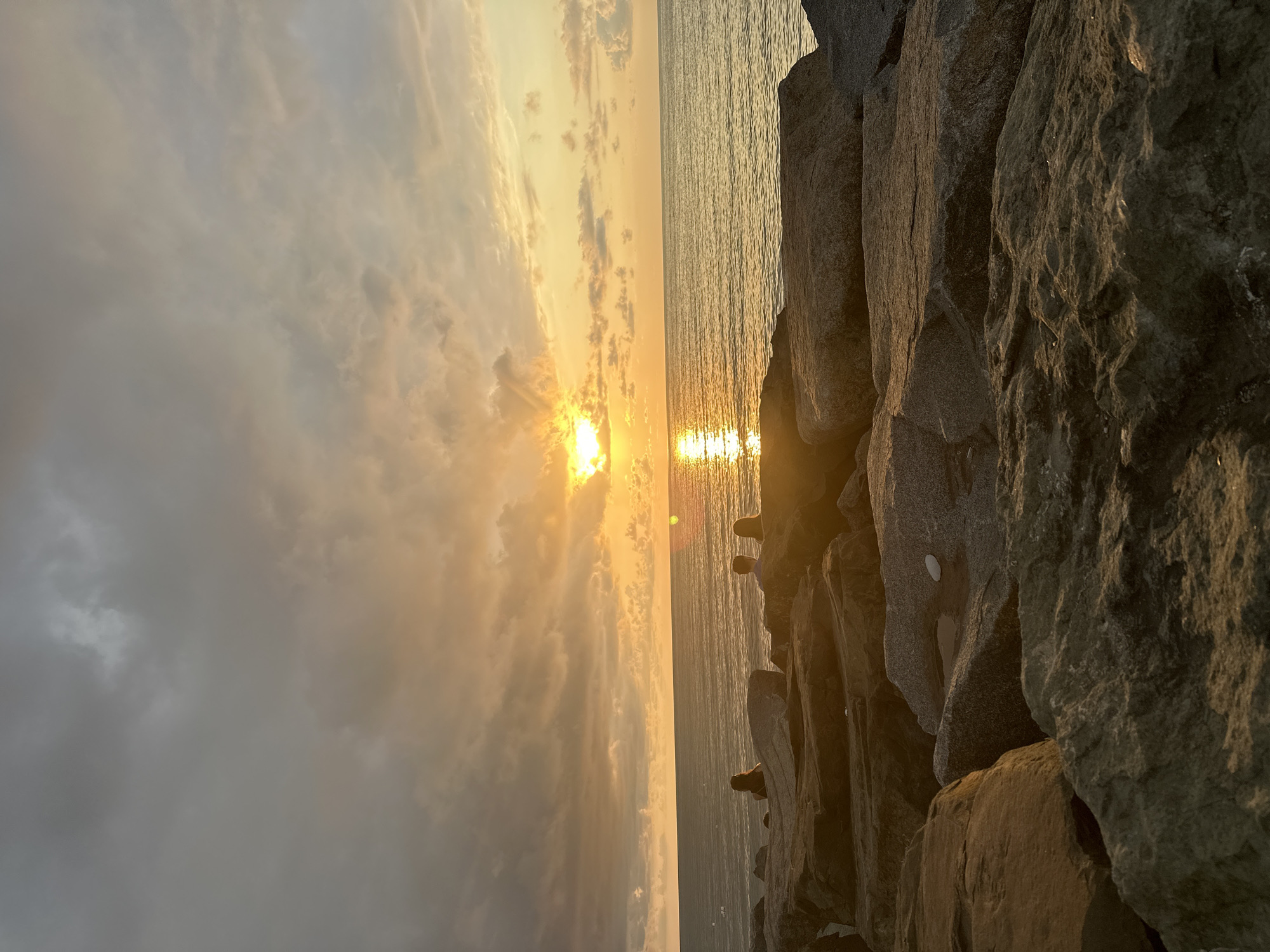 An image captures a serene sunset over the Black Sea, likely on the Batumi Boulevard in Georgia. The sky is a canvas of dramatic clouds, tinged with hues of orange and gold as the sun dips below the horizon. Rays of light pierce through the clouds, casting a shimmering reflection on the calm water. In the foreground, large, weathered rocks form a natural embankment, providing a vantage point for several silhouetted figures. Three individuals are visible, seated on the rocks, gazing out at the sea and the fading light. The overall scene evokes a sense of tranquility and natural beauty.