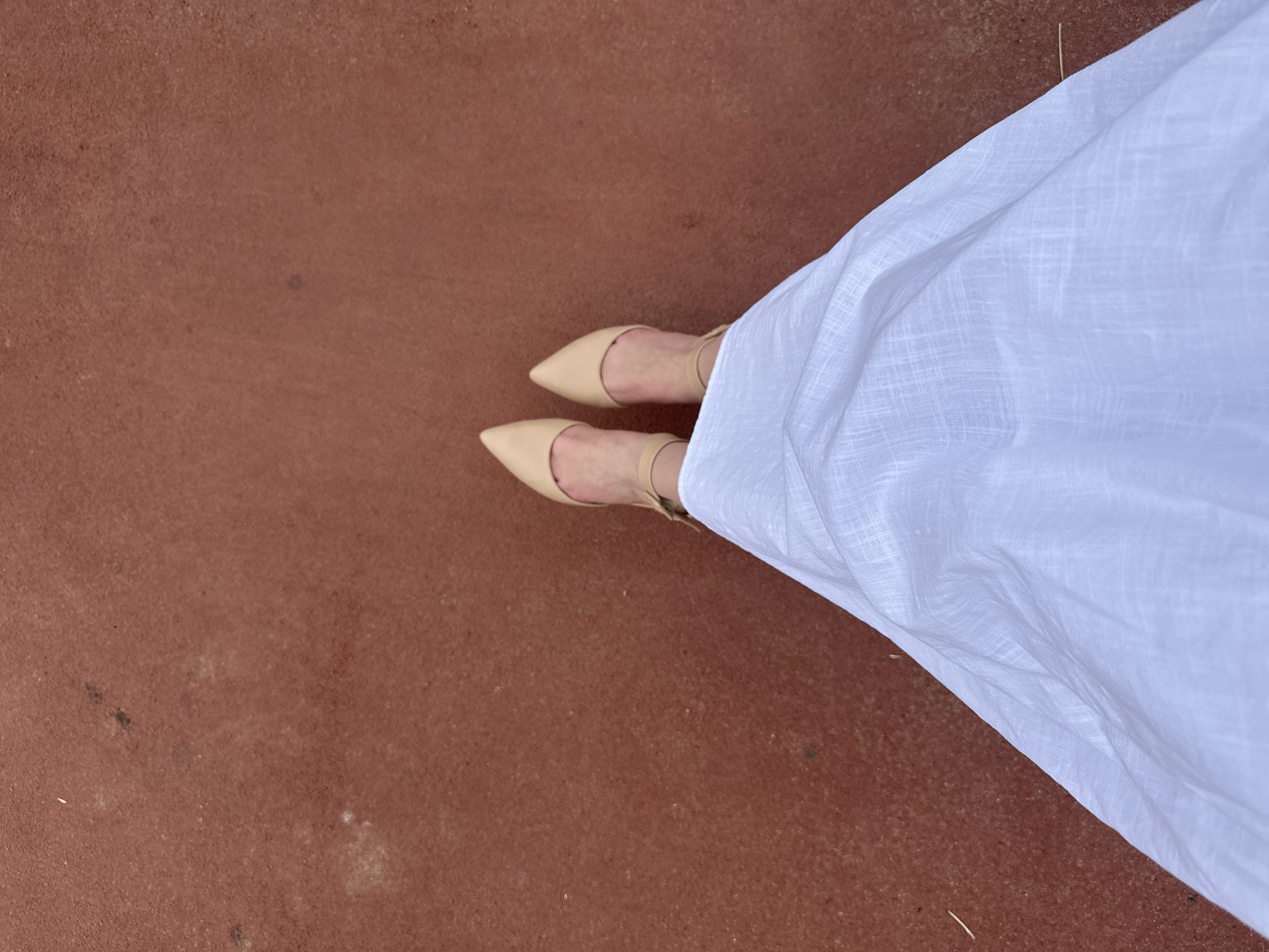 This media file captures a top-down view of a person's feet and legs, standing on a reddish-brown surface that appears to be outdoor pavement or a similar textured ground. The individual is wearing light-colored, pointed-toe flats with thin straps around the ankles. A flowing white garment, possibly a dress or skirt, drapes over the right leg and extends towards the bottom right of the frame, creating a contrast with the darker ground. The fabric of the white garment shows a subtle woven pattern. The lighting suggests it is daytime, with no clear indicators of time of day or weather conditions. The scene is minimalist, focusing on the fashion elements and the texture of the ground. There is no visible text in the image.