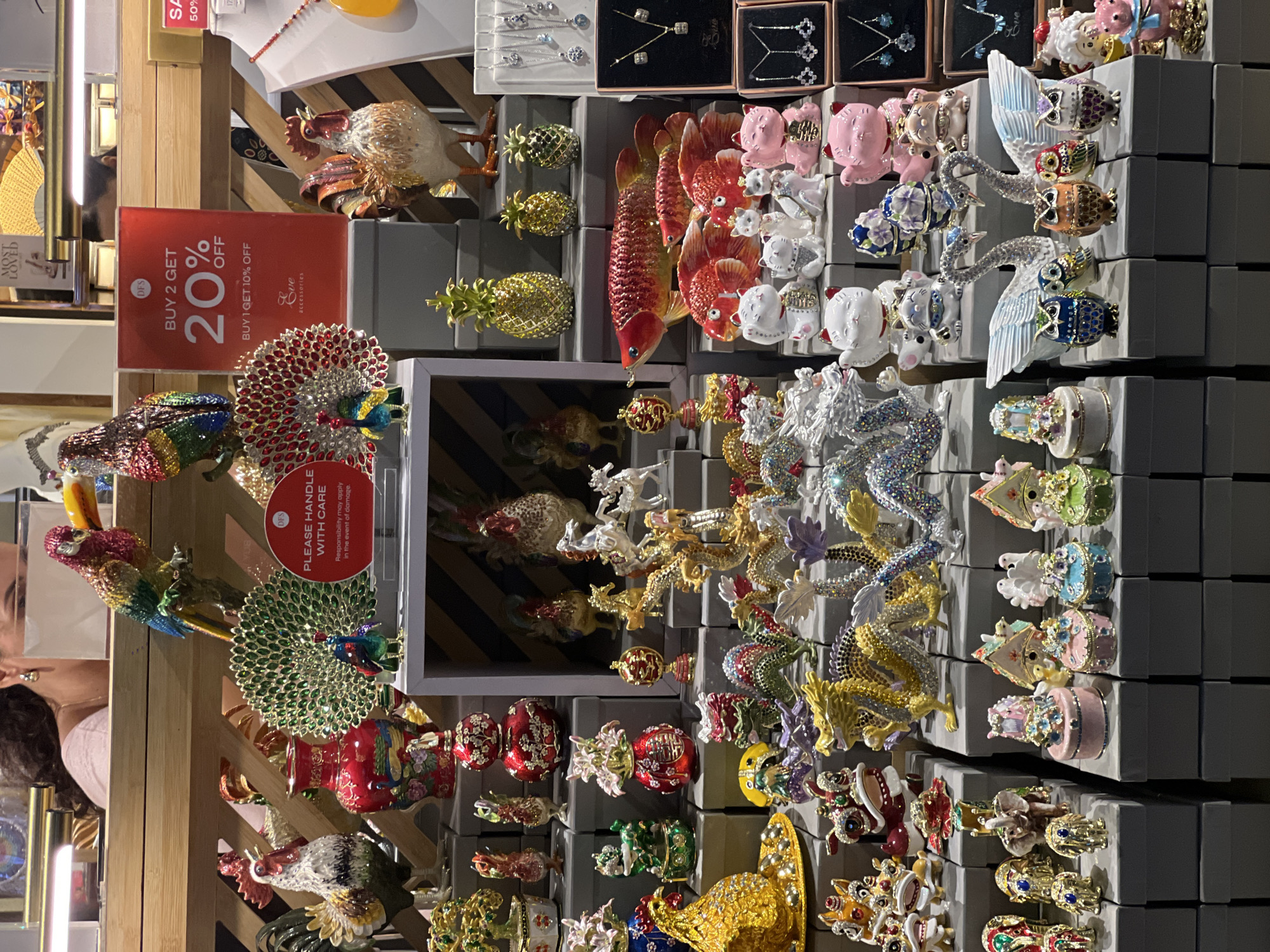 This image presents a vibrant and densely packed retail display, likely in a gift shop or accessory store in Kuta, Indonesia. The primary subjects are numerous ornate, colorful trinket boxes and figurines, crafted from metal with intricate enamel work and embedded rhinestones or crystals. These decorative items depict a wide array of animals and objects, including large peacocks with fan-like tails, majestic roosters, shimmering goldfish, golden pineapples, powerful dragons, graceful swans, lucky cats, elephants, and whimsical miniature cakes.
The merchandise is arranged on tiered shelves and individual gray boxes, creating a rich visual tapestry. Some jewelry, such as necklaces and earrings, is also visible in smaller boxes at the top. A person is partially visible on the left side of the frame, appearing to be browsing the selection.
Several pieces of text are visible on prominent signs. A large red sign advertises a sale, stating "DFS BUY 2 GET 20% OFF" and "BUY 1 GET 10% OFF," along with "Eve accessories." Another red sign carries a warning: "PLEASE HANDLE WITH CARE Responsibility may apply in the event of damage." Other partially visible texts include "DFS," "SA 50%", "MOST LOVED", and "100". The scene suggests an indoor setting with artificial lighting, focused on showcasing a collection of delicate and visually striking collectibles or souvenirs.