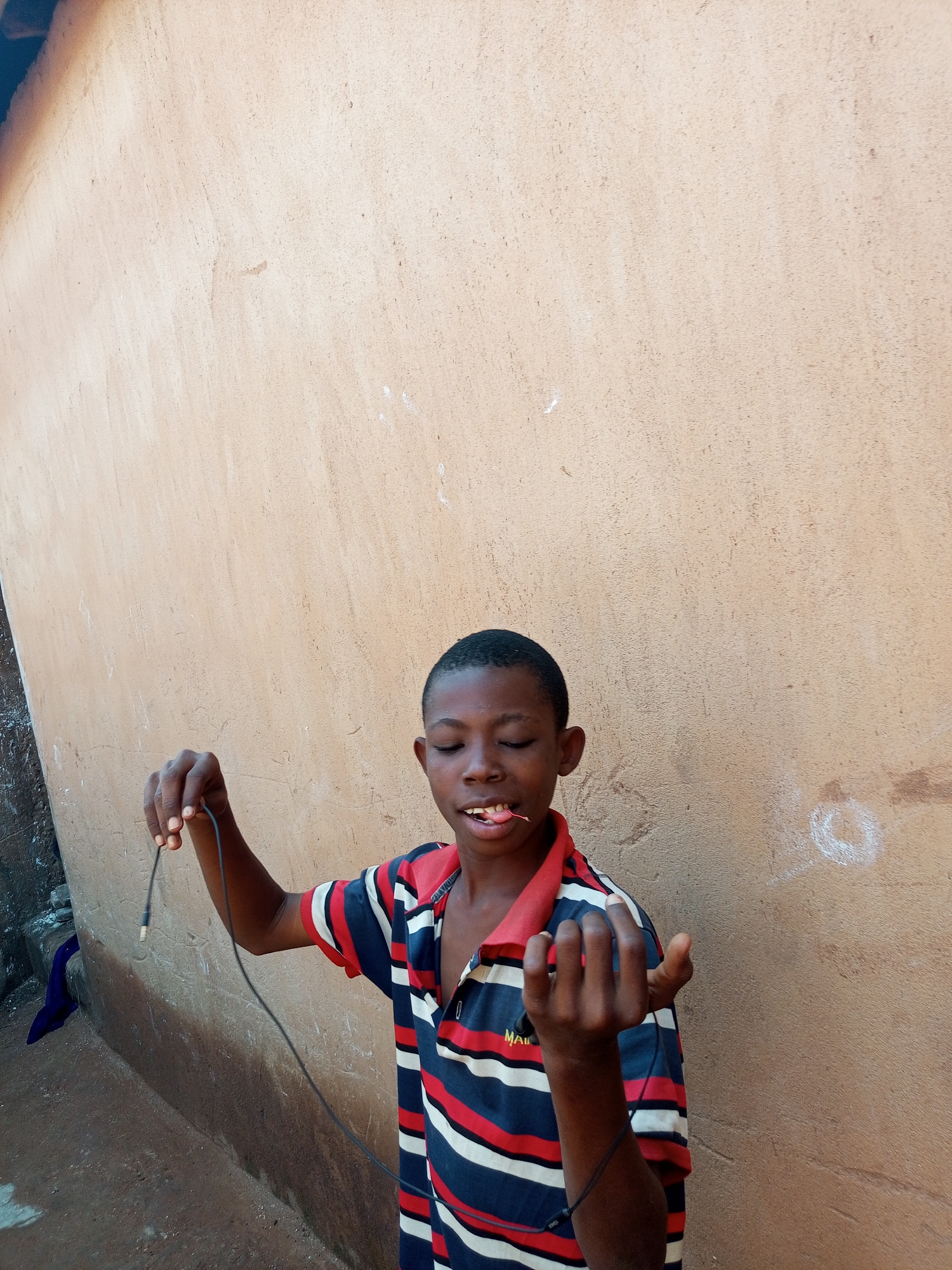 A young boy, with his eyes closed and a pink object protruding from his mouth, stands in front of a textured, light brown wall. He is wearing a striped polo shirt with navy, red, and white bands. In his right hand, he holds a black wire, which appears to be connected to an earpiece. The boy's left hand is open, with his fingers extended. The background suggests an outdoor setting, possibly a street in Tempure, Nigeria, given the presence of a rough, earth-toned wall and the casual attire of the boy. The lighting is natural and appears to be from an overhead source.