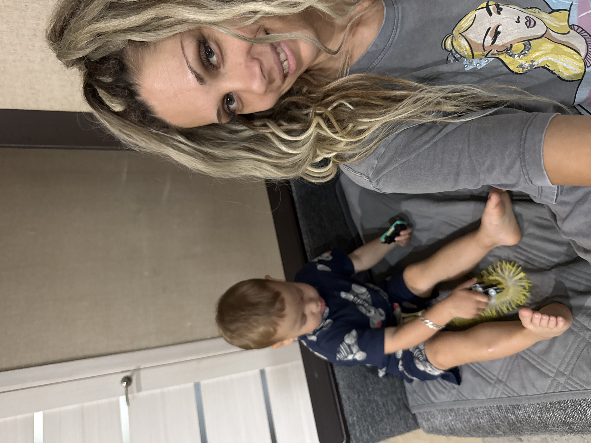 In Rusanovo, Russia, a close-up selfie captures a woman with blonde, curly hair pulled into a high ponytail, wearing a grey t-shirt with a cartoon graphic of a woman. Her right arm is visible, with her hand resting near her face. To her left, a young child sits on a grey quilted surface, wearing a dark blue t-shirt with a white butterfly pattern. The child's legs are visible, and they are holding a green toy car and a yellow fuzzy toy. The background appears to be an indoor setting with a textured wall and a white door with horizontal panels and a silver handle.