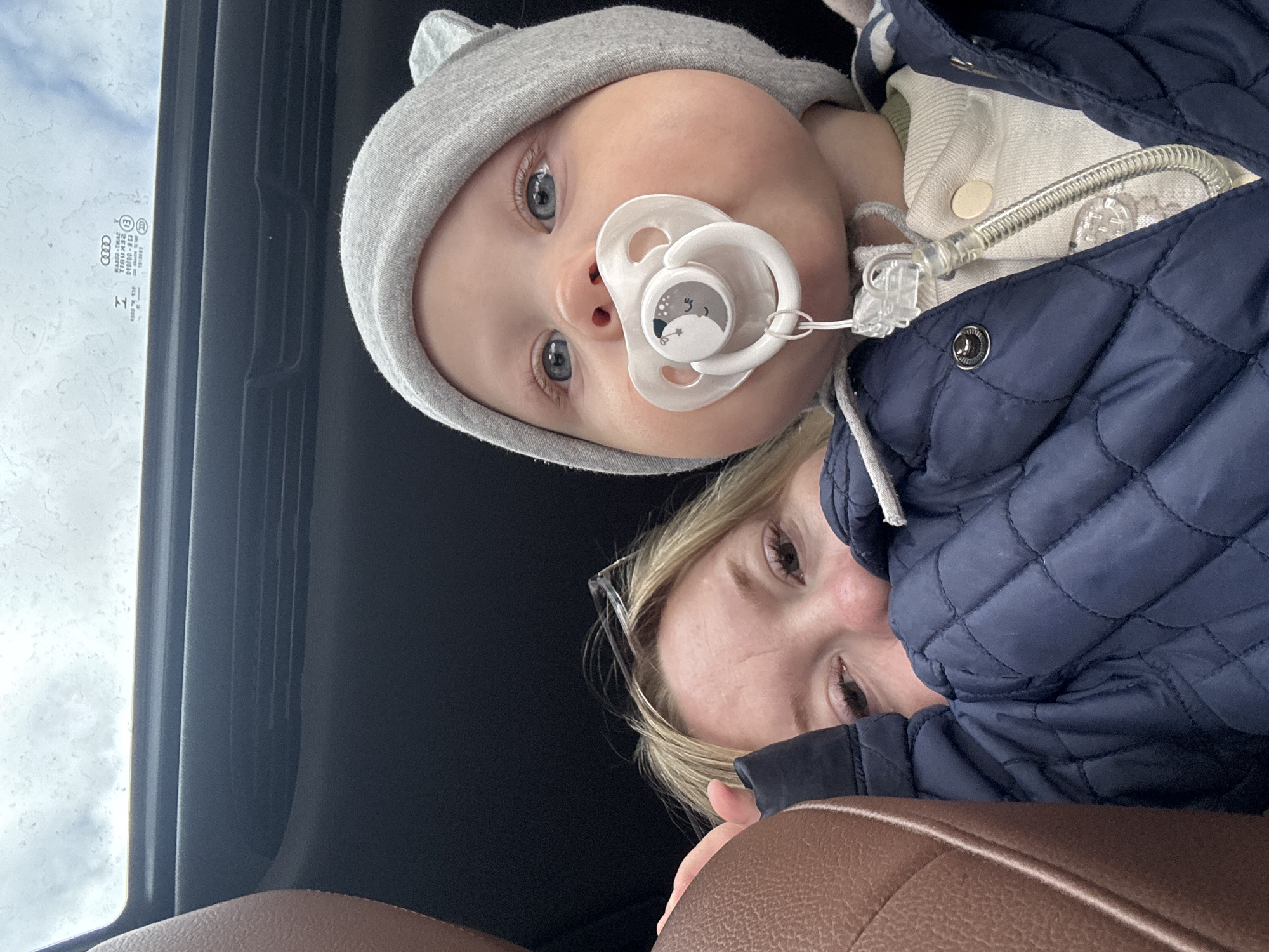 A photo taken inside a vehicle shows a baby with blue eyes and a gray hat with bear ears is wearing a pacifier and a blue jacket over a white outfit. The baby is positioned closer to the camera than an adult female with blonde hair who is also visible. The interior of the vehicle includes a dark ceiling and a brown seat. An Audi windshield sticker is visible near the top left of the image. There is no information to indicate the location of this scene is in Chaikovsky, Russia.