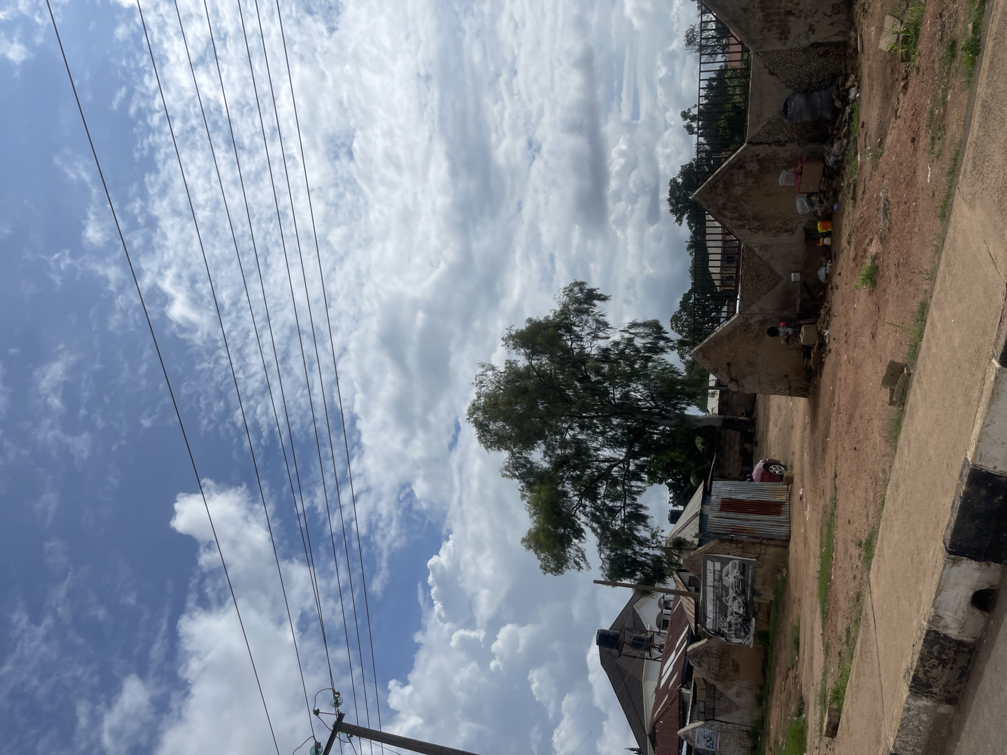 In Bar Jirgi Summa, Nigeria, a scene unfolds under a partly cloudy sky. Power lines crisscross the frame, suspended from a utility pole on the left. In the middle ground, a large tree with sparse foliage stands tall. To the left of the tree, a multi-story building with a tiled roof and water tanks is visible. In front of the tree, a small, corrugated metal shed with a red door is positioned. A red car is parked nearby. The foreground features a dirt road with a concrete curb. Several rudimentary structures with triangular roofs and rough, plastered walls are scattered across the landscape. One of these structures has a sign advertising an "Automotive Workshop" with images of cars. Debris and various items, including buckets and a large trash bag, are present near the structures. A metal fence is visible in the background, separating the area from more distant buildings and trees.