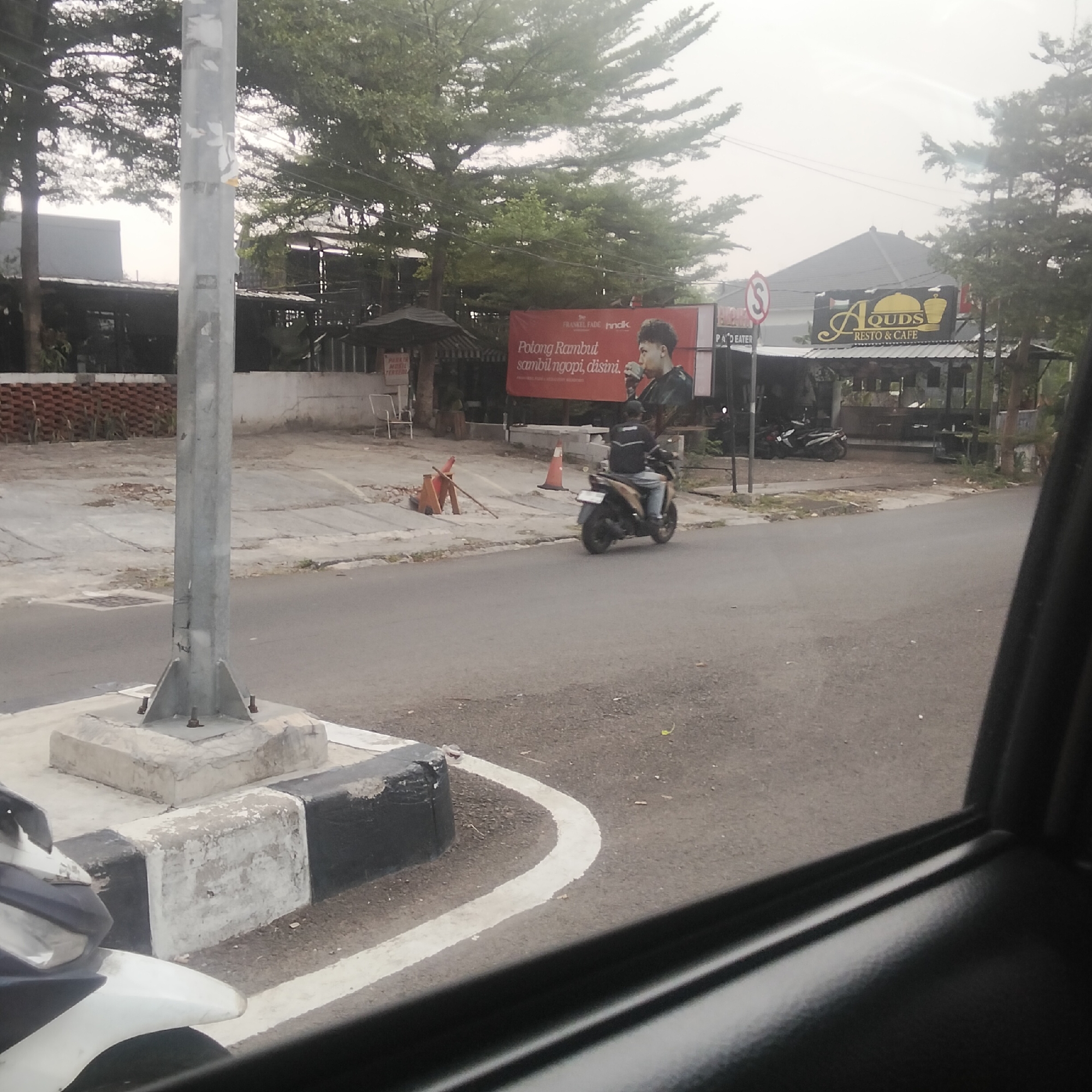 The image depicts a street scene with a man riding a motorcycle. The man is wearing a dark jacket and jeans and is looking forward. The motorcycle is dark in color and has a license plate. The street is paved and there is a sidewalk on the right side of the image. There are trees and buildings in the background. A sign for "AOUDS RESTO & CAFE" is visible on the right side of the image. A red banner with Indonesian text that translates to "Cut Hair while Coffee" is also visible. The scene appears to be located in a commercial area, possibly in the city of Bogor, Indonesia.