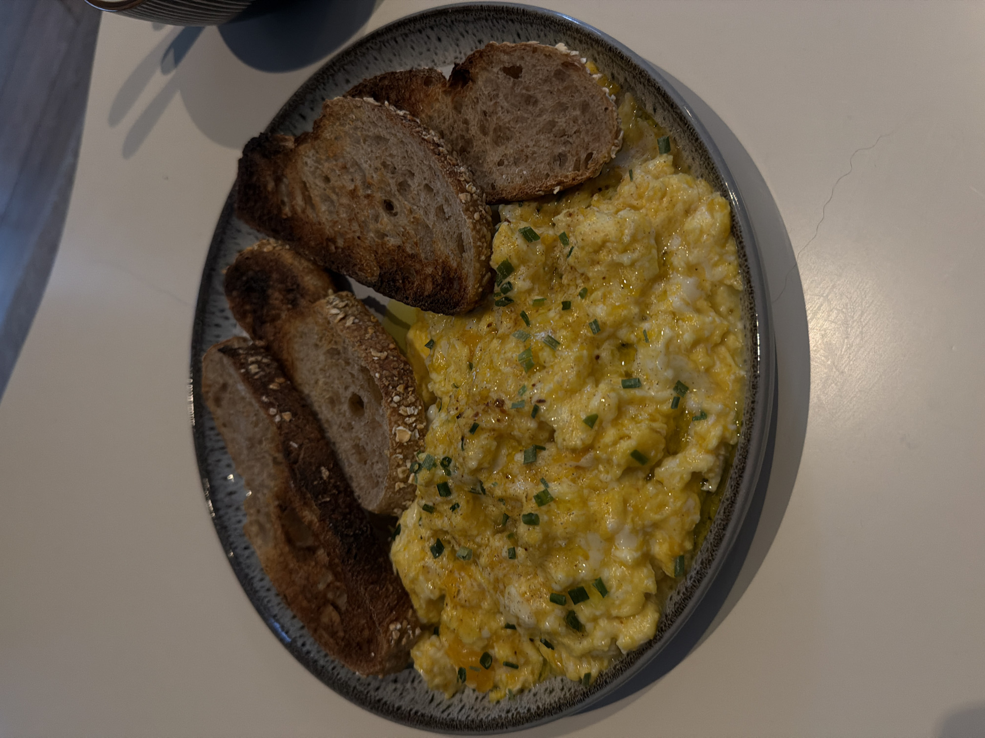A close-up, overhead shot captures a plate of scrambled eggs and toast. The plate, dark gray with a textured rim, sits on a light-colored surface, possibly a table. The scrambled eggs are a vibrant yellow, interspersed with what appear to be finely chopped green chives, suggesting a garnish. They are glistening, indicating they may have been cooked with butter or oil. Arranged around the eggs are several slices of toasted bread, each showing a golden-brown crust and a porous interior. The toast appears to be whole grain, with visible seeds or oats. The lighting creates soft shadows on the table, suggesting an indoor setting. The overall impression is a simple, yet appealing breakfast or brunch dish. There is no discernible information in the image to suggest a specific location in London, United Kingdom.
