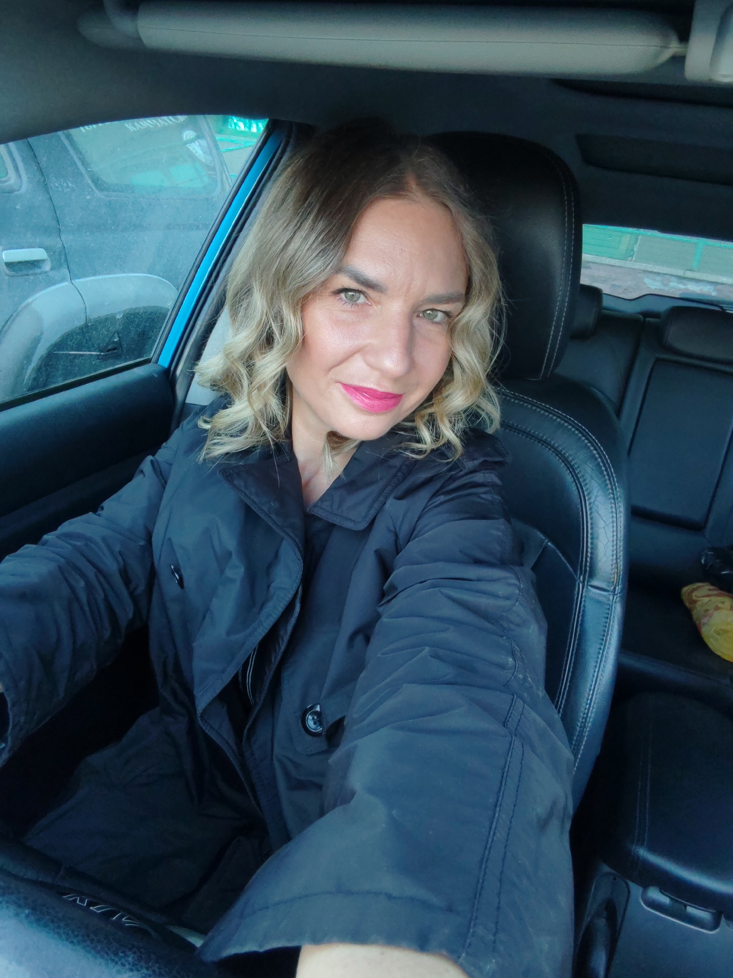 A woman with blond, curly hair and bright pink lipstick sits in the driver's seat of a car. She is wearing a black coat. The car appears to be parked, as another vehicle is visible through the driver's side window. The interior of the car is primarily black, with leather seats. It is not possible to determine the specific location in Petropavlovsk-Kamchatsky, Russia, from the visual information provided.