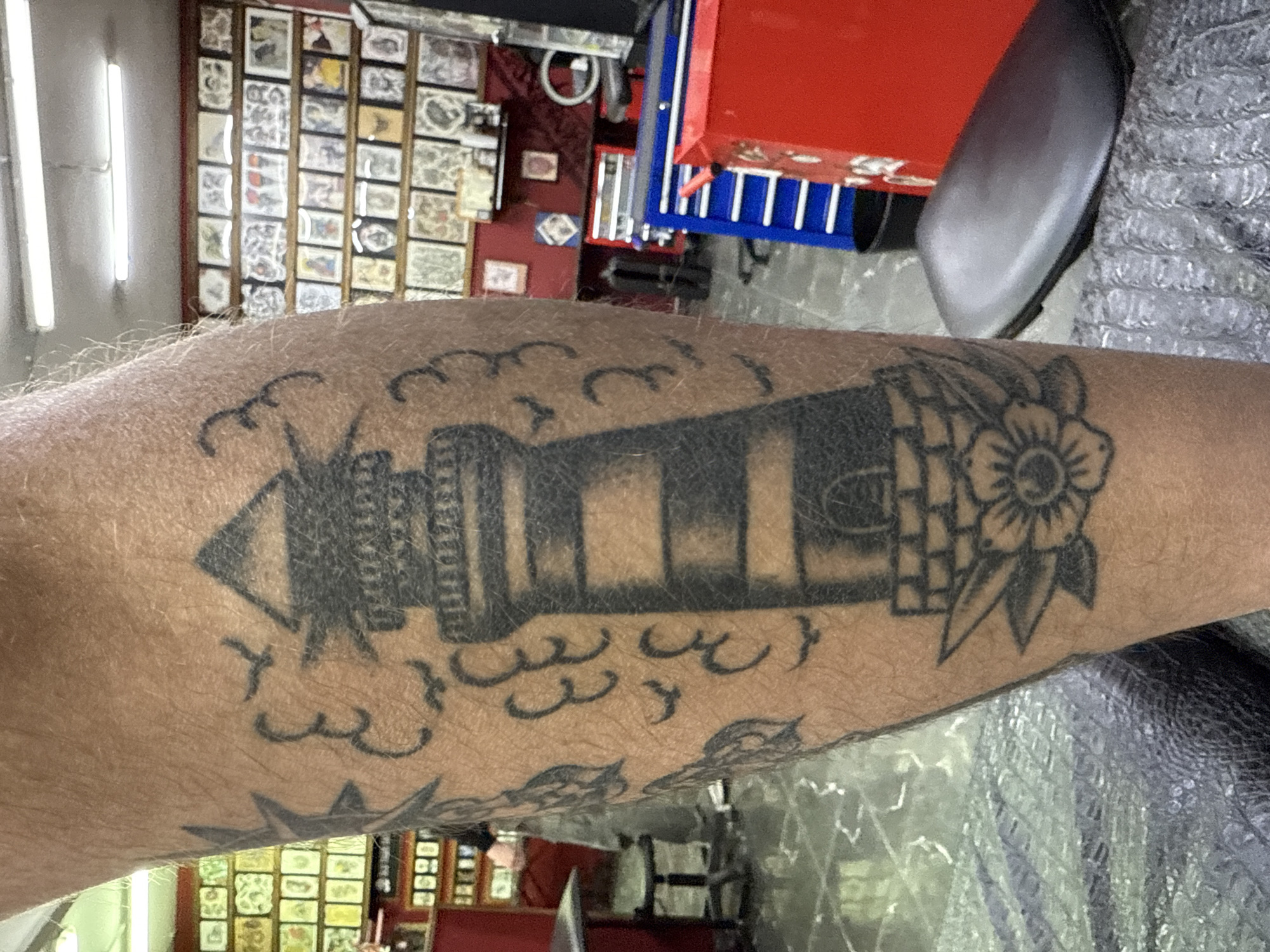 The image depicts a close-up of a forearm with a black and grey lighthouse tattoo. The tattoo features a traditional style lighthouse with a flower on one side and cloud-like shapes around it. In the background, a wall covered in framed artwork is visible, possibly indicating the setting is a tattoo parlor. A red and blue tool chest is also visible. The location is undetermined, and Moscow, Russia, cannot be confirmed from the image. There are no people visible.