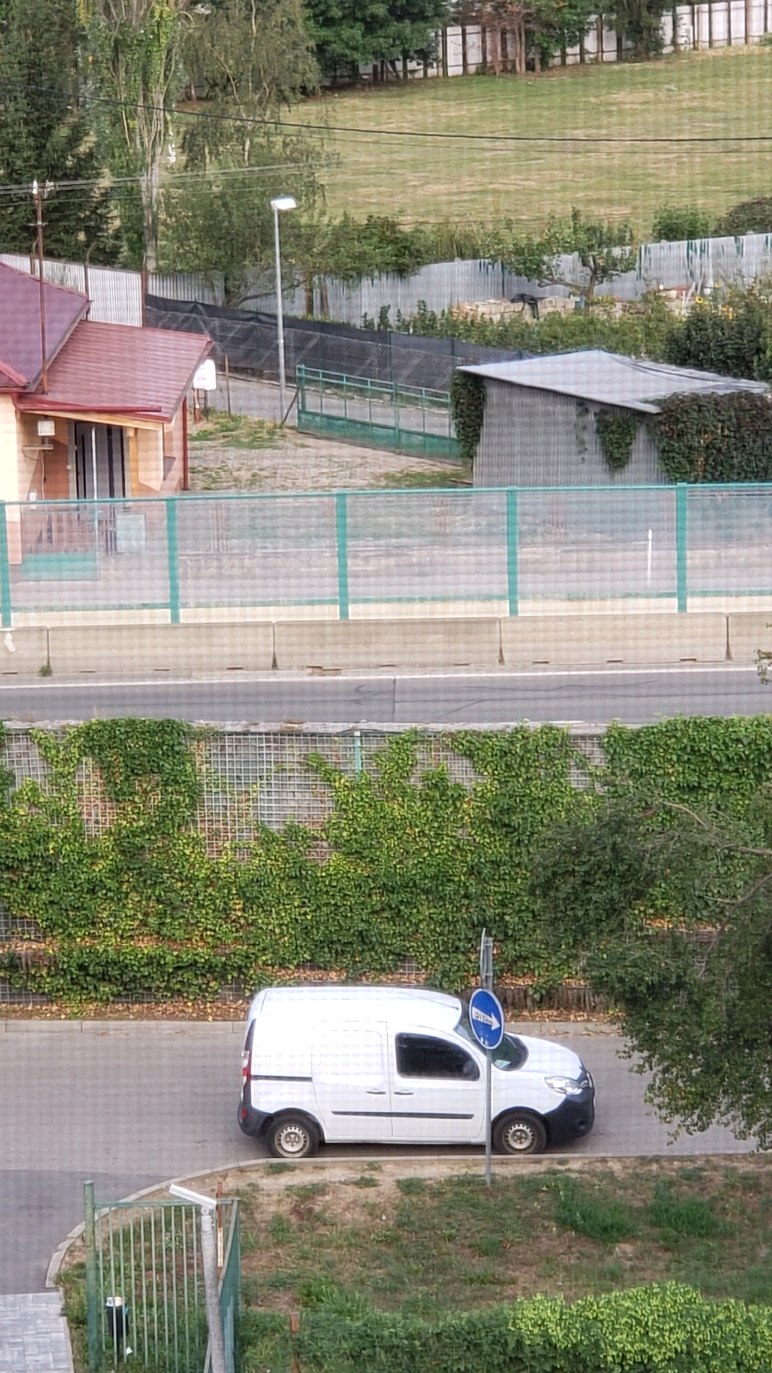 The image captures a street scene in Prešov, Slovakia, with a white panel van parked on the right side of the road. The van is a modern model, likely used for commercial purposes, and appears to be stationary. It is parked adjacent to a wall covered in dense green ivy, which also forms a backdrop to the scene. A blue circular traffic sign with a white arrow indicating a right turn is visible next to the van. The road surface is asphalt, and a concrete barrier separates it from a grassy area with scattered trees and bushes. In the background, a light-colored building with a red roof is partially visible, along with a metal fence and a street lamp. The overall impression is one of an ordinary urban environment on a clear day.
