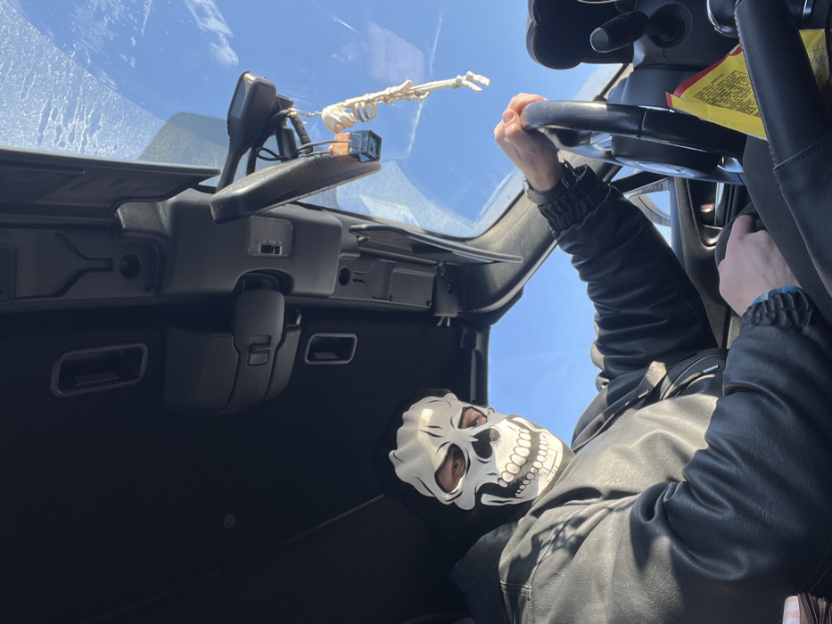 An individual is depicted seated in the driver's position inside a vehicle, located in Saint Petersburg, Russia. The person is wearing a white and black skull-patterned mask, covering their face with their eyes visible through the eyeholes, and a dark, possibly black, hooded jacket. Their left hand, wearing a wristwatch, and their right hand, wearing a blue bracelet, are gripping the black steering wheel. The vehicle's interior features a black headliner and overhead console with integrated compartments. A rearview mirror is centrally positioned, from which a small, light-colored skeleton figurine and a dark, rectangular charm are suspended. A clear, blue sky is visible through the windshield, with minor frosty patterns or reflections on the upper glass. A yellow paper object is visible on the passenger side, near the center console. The individual is looking directly forward towards the camera.