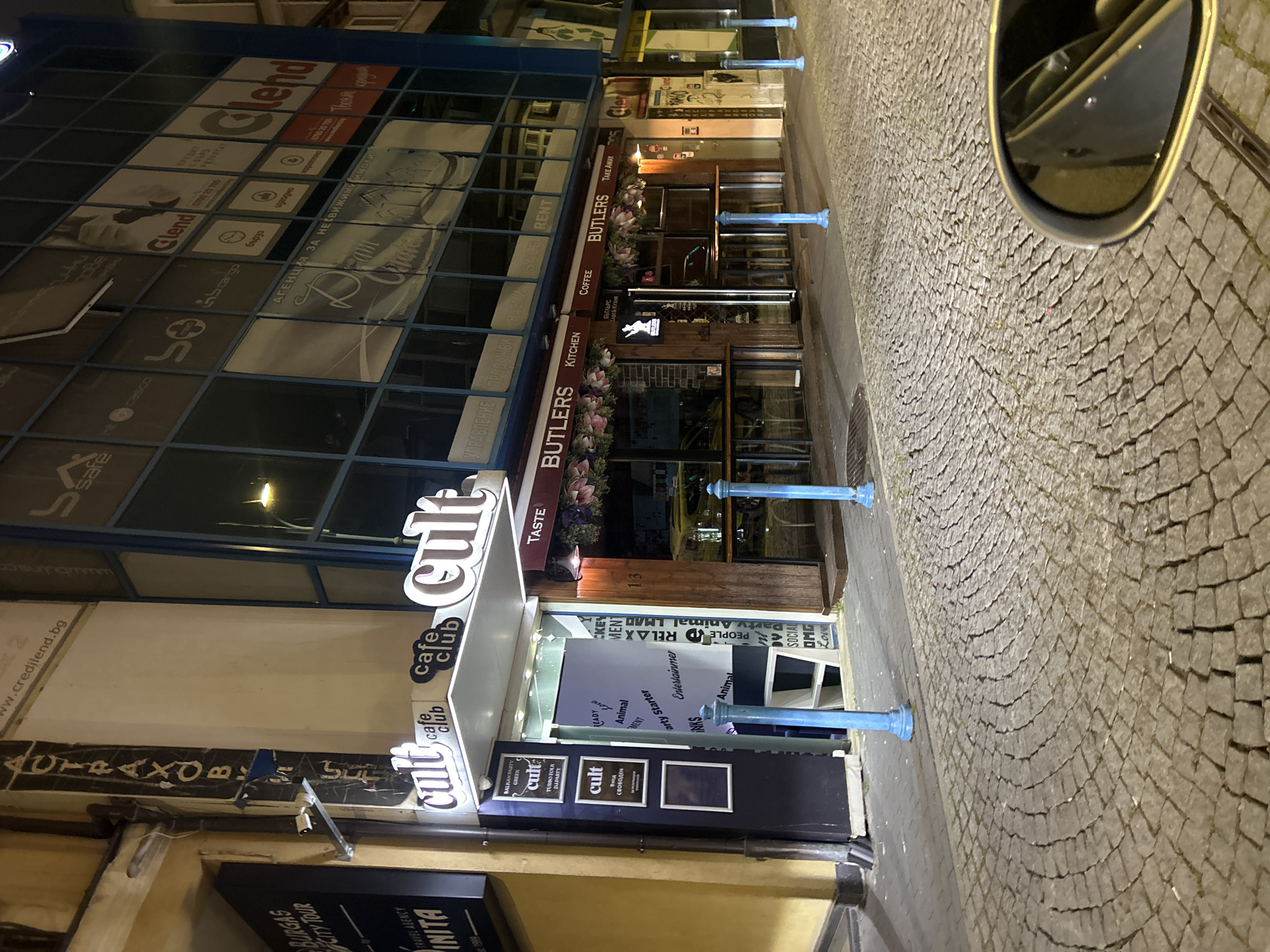 This is an exterior shot of a street scene in Burgas, Bulgaria, showing the fronts of two businesses and a cobblestone street. “Butlers” a restaurant or cafe with the signs “Kitchen,” “Coffee,” and “Take Away” above the entrance. Next to it is “Cult Cafe Club.” A sign beneath the cafe reads “Burgas City Tour Travel Agency INITIA.” The adjacent building has large windows with several different logos in each window, including “Clend.” Blue bollards line the street. A side mirror from a vehicle is in the top-right.