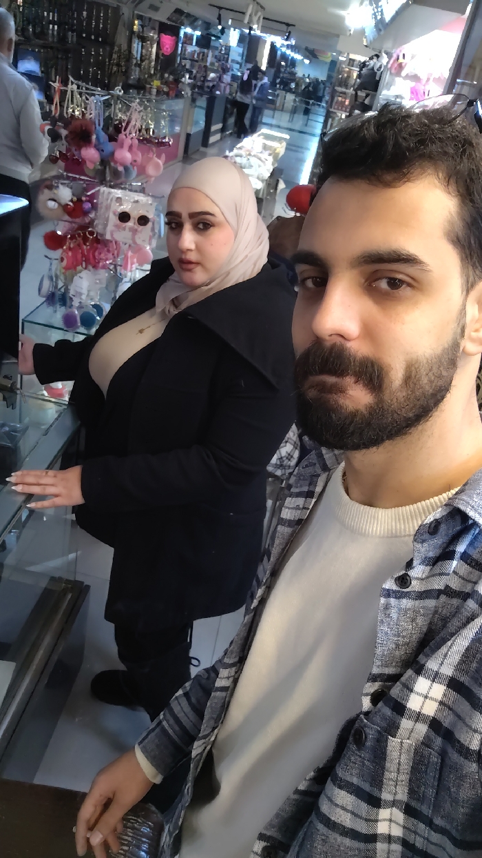 This image depicts an indoor scene within a retail establishment, likely a shopping mall or store, located in Barza Municipality, Syria. In the foreground, a male individual with dark hair, a beard, and a mustache is seen, appearing to take a selfie. He is wearing a white crew-neck shirt under an open blue and white plaid long-sleeve shirt. His right hand is visible in the lower-left corner, seemingly holding the camera device.
Behind him and to the left, a female individual is positioned. She wears a light beige hijab, a dark-colored (likely black) jacket with a wide collar, and a light-colored top beneath. Her left hand, featuring light-colored manicured nails, rests on the surface of a glass display case. She is looking towards the camera.
The background shows multiple glass display cases filled with various consumer goods, including colorful keychains, sunglasses, and other accessories. Additional blurred figures are visible further in the background, suggesting other patrons or staff within the establishment. The flooring appears light-colored and reflective, illuminated by overhead lighting fixtures typical of a commercial interior.