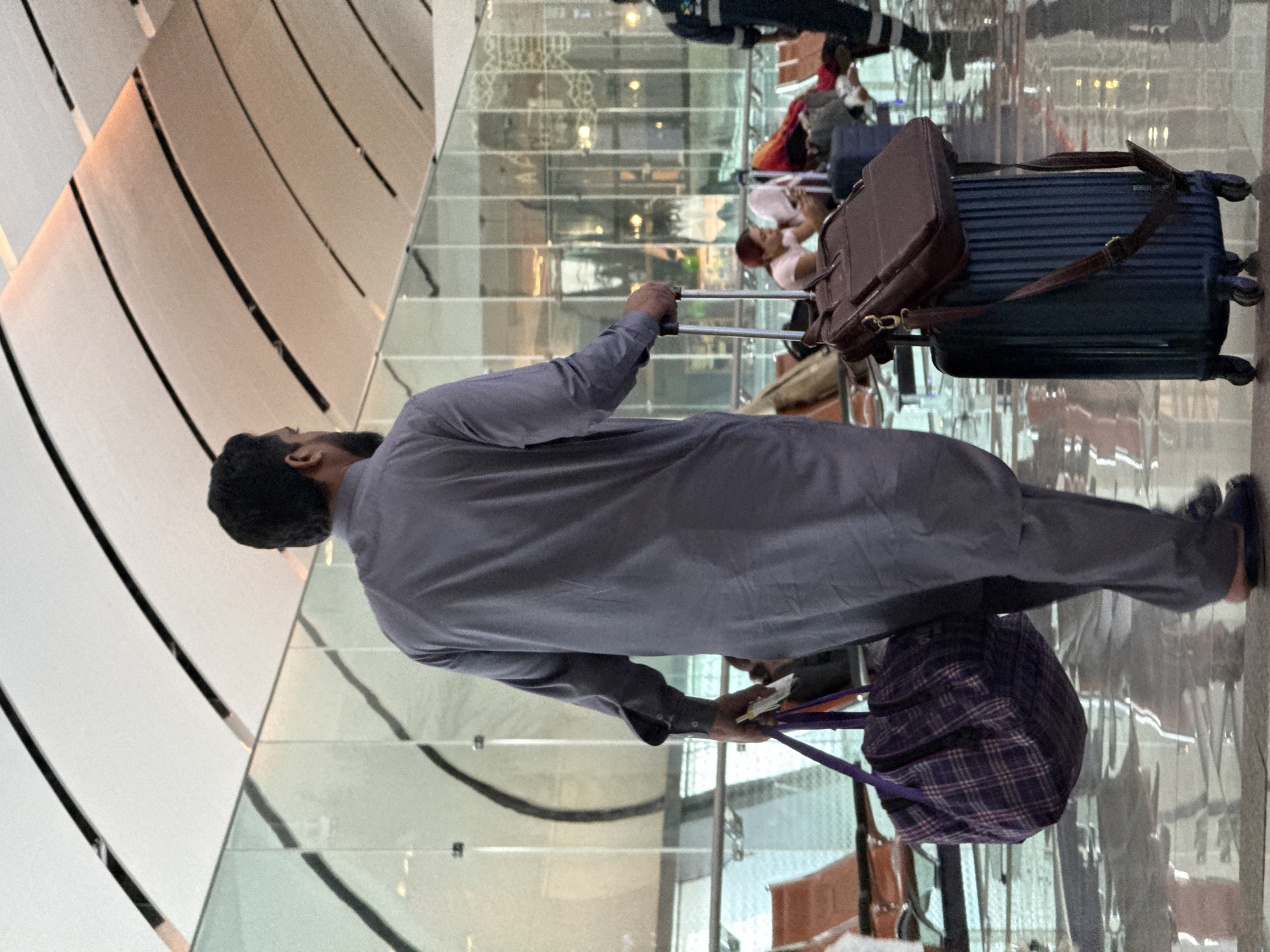 An adult male, facing away from the camera, walks through an airport terminal. He is wearing a grey shalwar kameez and black sandals. In his left hand, he carries a purple and white plaid duffel bag. In his right hand, he holds the handle of a dark blue wheeled suitcase, topped with a dark brown leather briefcase. The airport features a modern architectural design with a high, curved ceiling and large glass windows. Orange seating areas are visible in the background, with other travelers seated. The floor is highly reflective, indicating a polished surface. The image appears to be taken in the Seeb Province of Oman, likely at Muscat International Airport, given the context of an airport terminal in Oman.