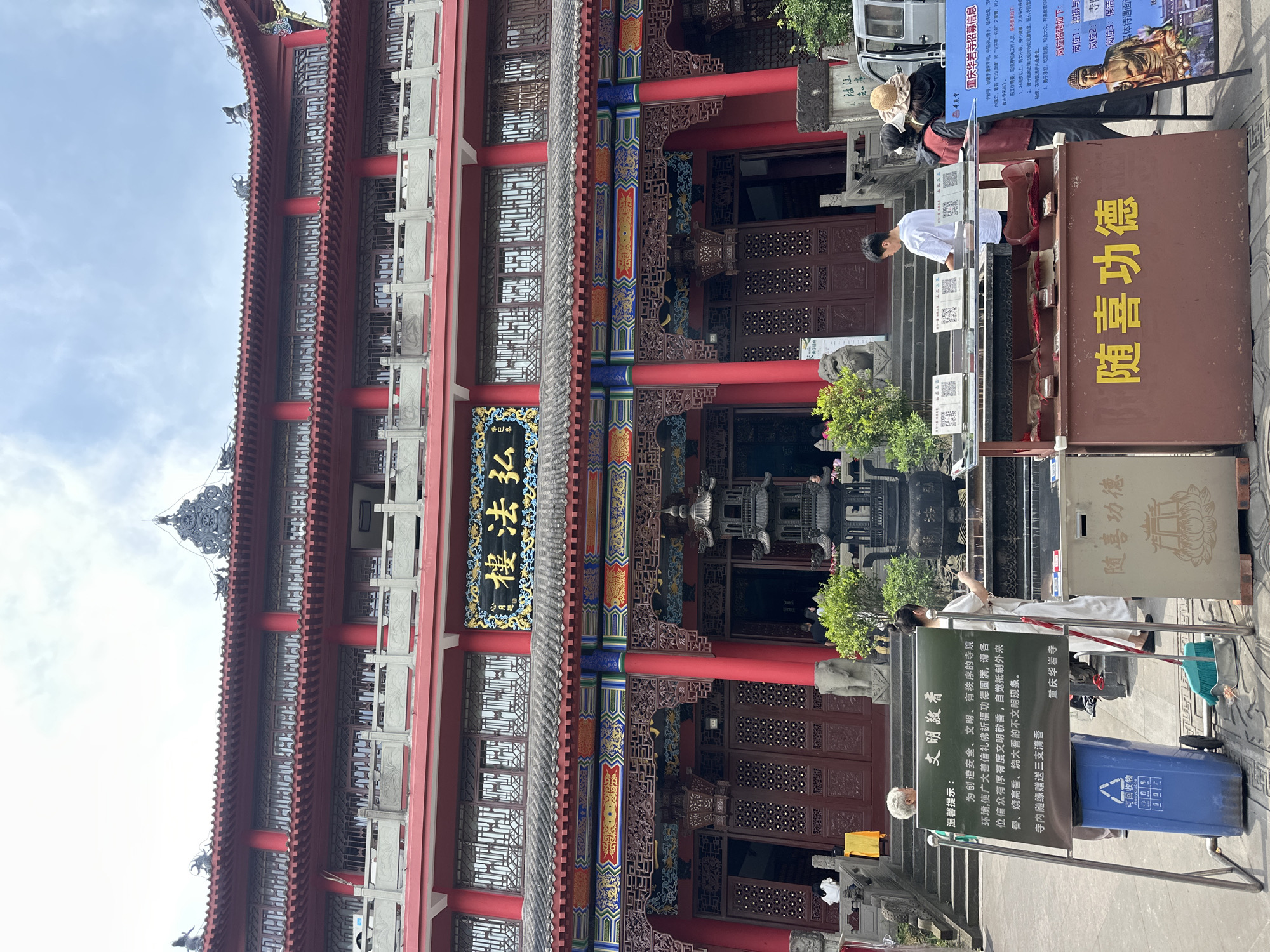 The image shows the exterior of a traditional Chinese temple in Jiulongpo District, China. The temple features multiple stories with red pillars, intricate woodwork, and traditional Chinese architectural details. There are people visible near the entrance, including some appearing to be worshippers. Signage in Chinese characters is displayed, one reading "随喜功德", which is related to giving donations. A blue recycle bin is visible. Additionally, there's a poster with information and an image of Buddha.