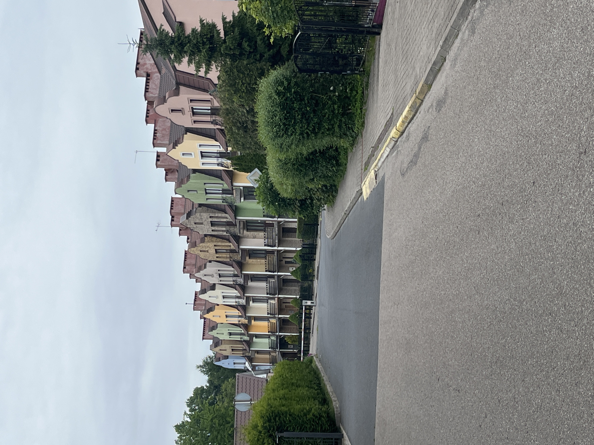 The image shows a row of colorful terraced houses with steep gabled roofs and chimneys, set against an overcast sky. A paved road curves to the left, with a sidewalk on the right. Several neatly trimmed hedges line the front of the houses and the edge of the sidewalk. A black metal gate and fence are visible to the right, behind a dense hedge. A red and white barrier arm is positioned across the road, suggesting a controlled access point. The houses are painted in various pastel shades, including pink, peach, yellow, light green, and blue. The architecture suggests a planned residential development. This scene is characteristic of the residential area of "Mayak" (Маяк) in Kaliningrad, Russia, known for its distinctive, brightly colored houses. There are no people visible in the image.
