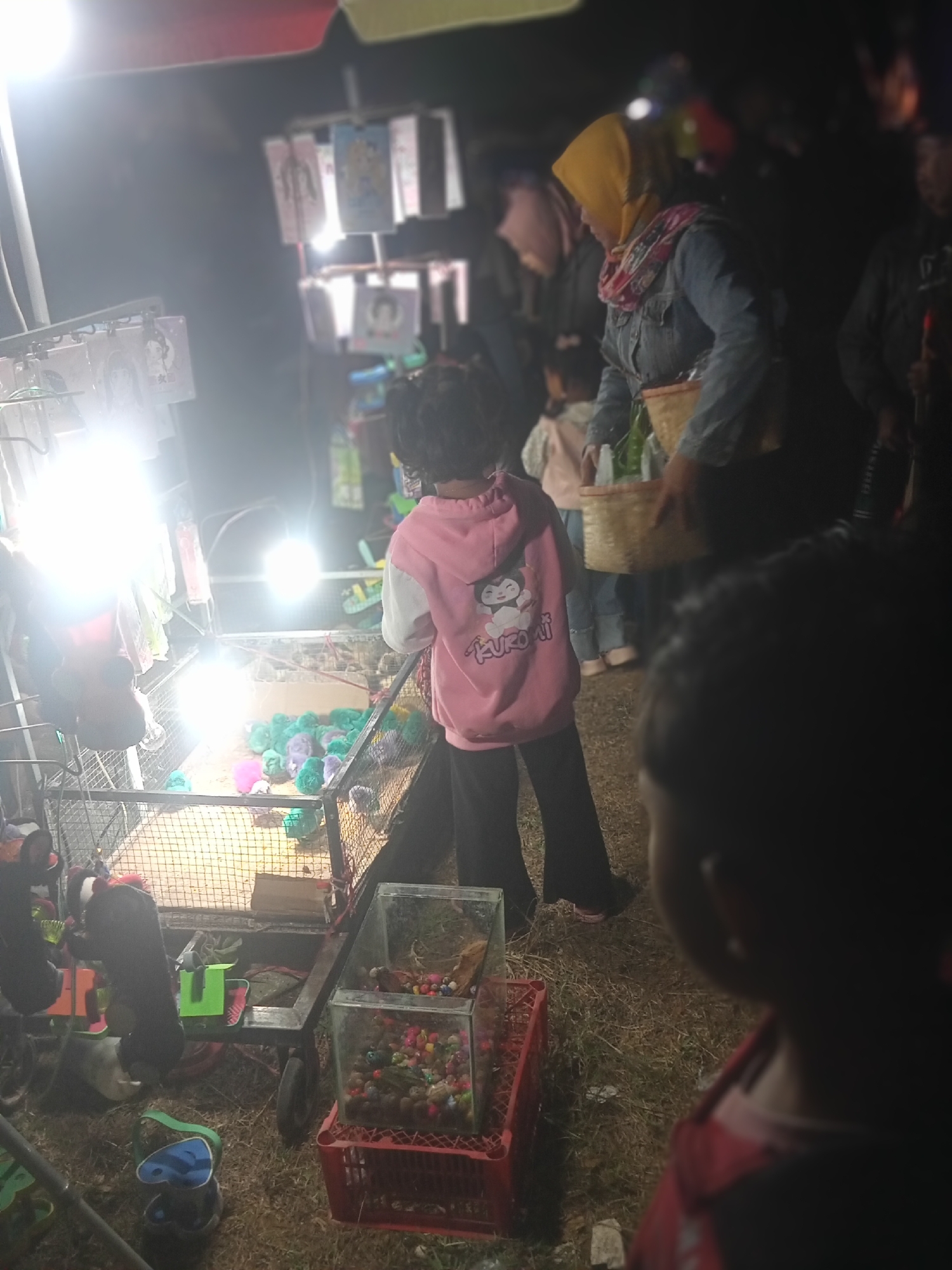 At night in Pule, Indonesia, a scene unfolds at what appears to be a roadside stall. A young child in a pink hooded sweatshirt, featuring a cartoon character and the word "Kuromi," stands facing a display of colorful objects in a wire cage. The child is positioned near the front of the image, with their back to the camera.
Behind the child, a woman wearing a yellow headscarf and a denim jacket is visible, holding a woven basket. She is interacting with the goods at the stall, with other people blurred in the background. The stall itself features various items hanging on racks, illuminated by bright lights that create glare. In the foreground, a small glass aquarium filled with colorful beads and stones sits on a red plastic crate. The ground is covered in grass and dirt. The overall atmosphere suggests a lively, informal marketplace or event.