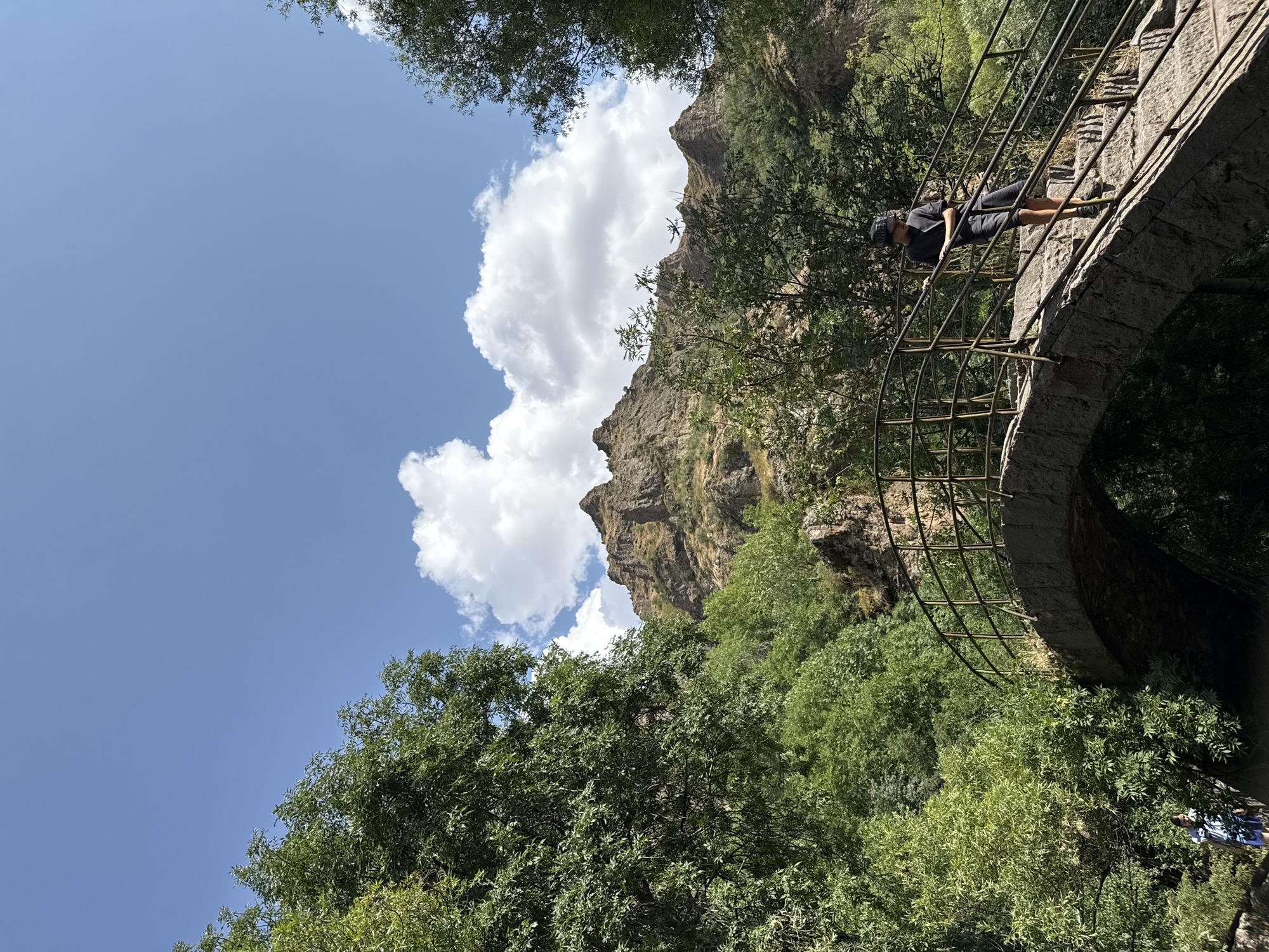 A person wearing a dark t-shirt and shorts, and a black baseball cap, walks down a stone bridge with metal railings in the city of Goght, Armenia. The bridge is arch-shaped and has weathered stone steps. Lush green trees surround the bridge, with a rocky, mountainous landscape visible in the background under a bright blue sky with fluffy white clouds. Another person in the distance, wearing a blue and white striped shirt and shorts, is partially visible near the base of the bridge.
