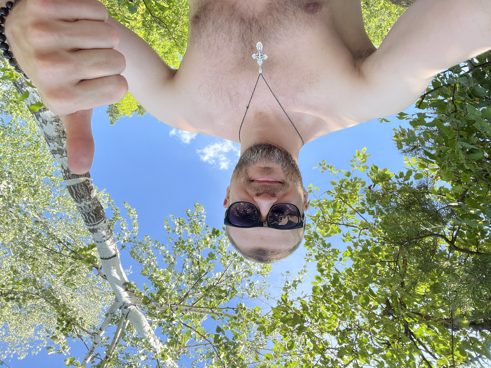 A shirtless man with a beard and sunglasses, wearing a cross necklace, gives a thumbs-up gesture. He is outdoors, surrounded by lush green trees and a bright blue sky with scattered clouds. The scene appears to be a sunny day in a natural setting, possibly a park or forest.