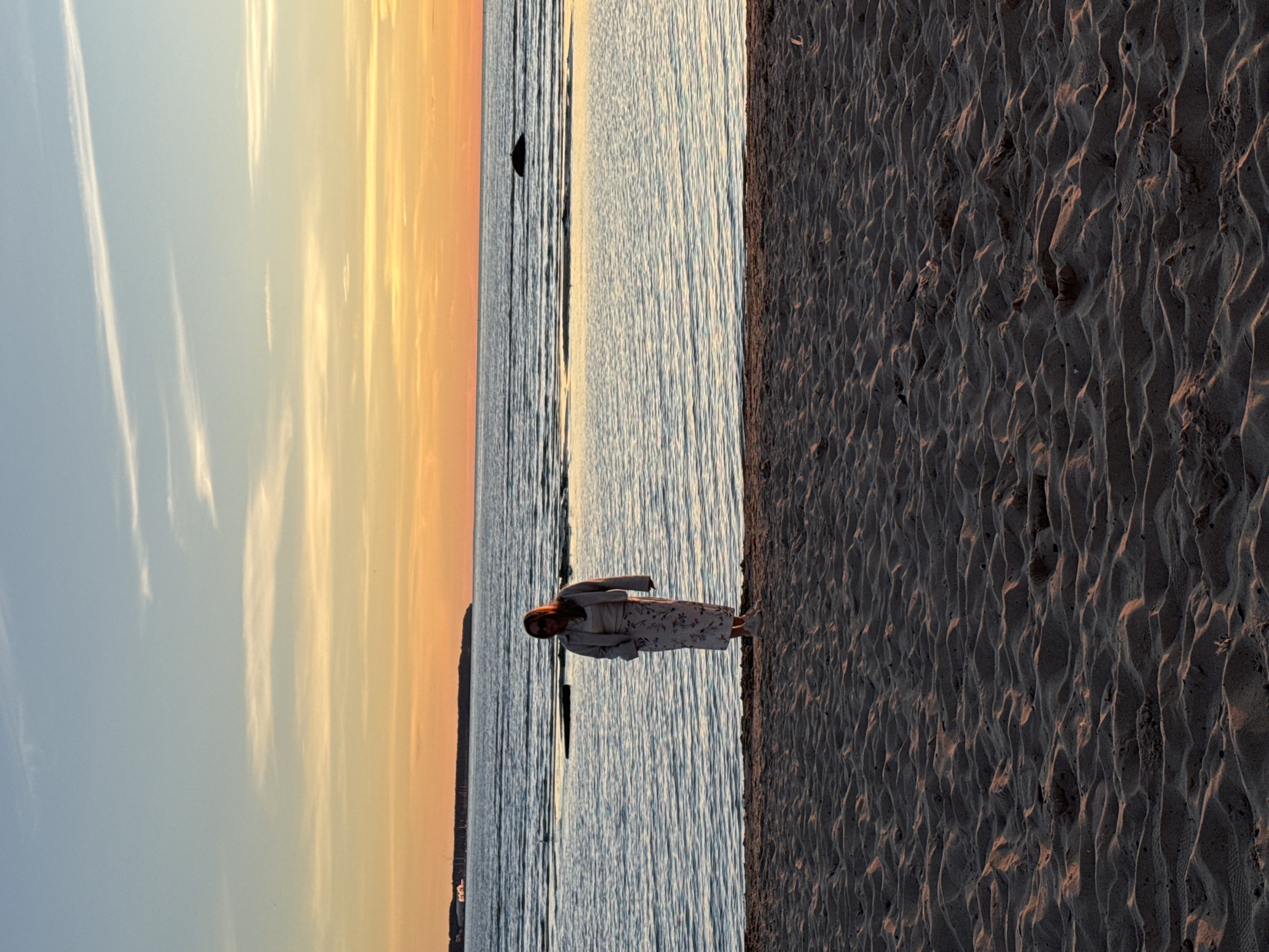 The image shows a woman standing on a sandy beach at sunset. The sky is a gradient of blue, orange, and yellow, with some wispy clouds. The sea is calm with gentle waves, and the sand is rippled from the wind. In the distance, a landmass with buildings and a dock can be seen, which is likely the city of Tallinn, Estonia. The woman is wearing a light-colored dress and a cardigan, and sunglasses. She is facing the viewer, and her hands are by her sides. The overall mood of the image is peaceful and serene.