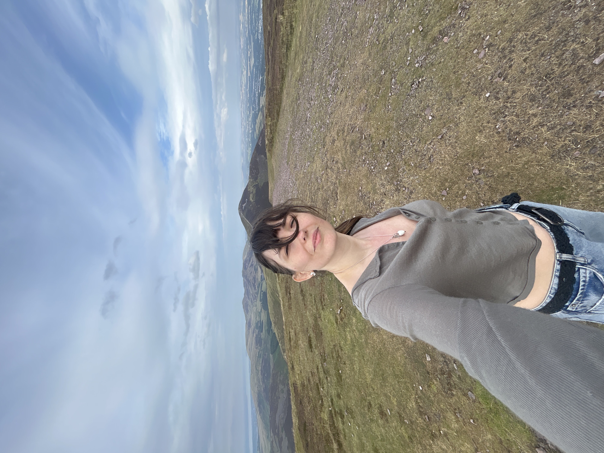 A woman is taking a selfie on a grassy hillside overlooking a valley with a town in the distance, likely near Penicuik, United Kingdom. The woman, with dark hair and bangs, wears a gray cropped shirt with buttons and blue jeans. She has a necklace with a white pendant and earrings. The landscape consists of rolling hills covered in dry grass and heather, with a dirt path leading away from the viewer. The sky is cloudy with patches of blue. The overall scene suggests an outdoor recreational activity in a rural setting close to an urban area.