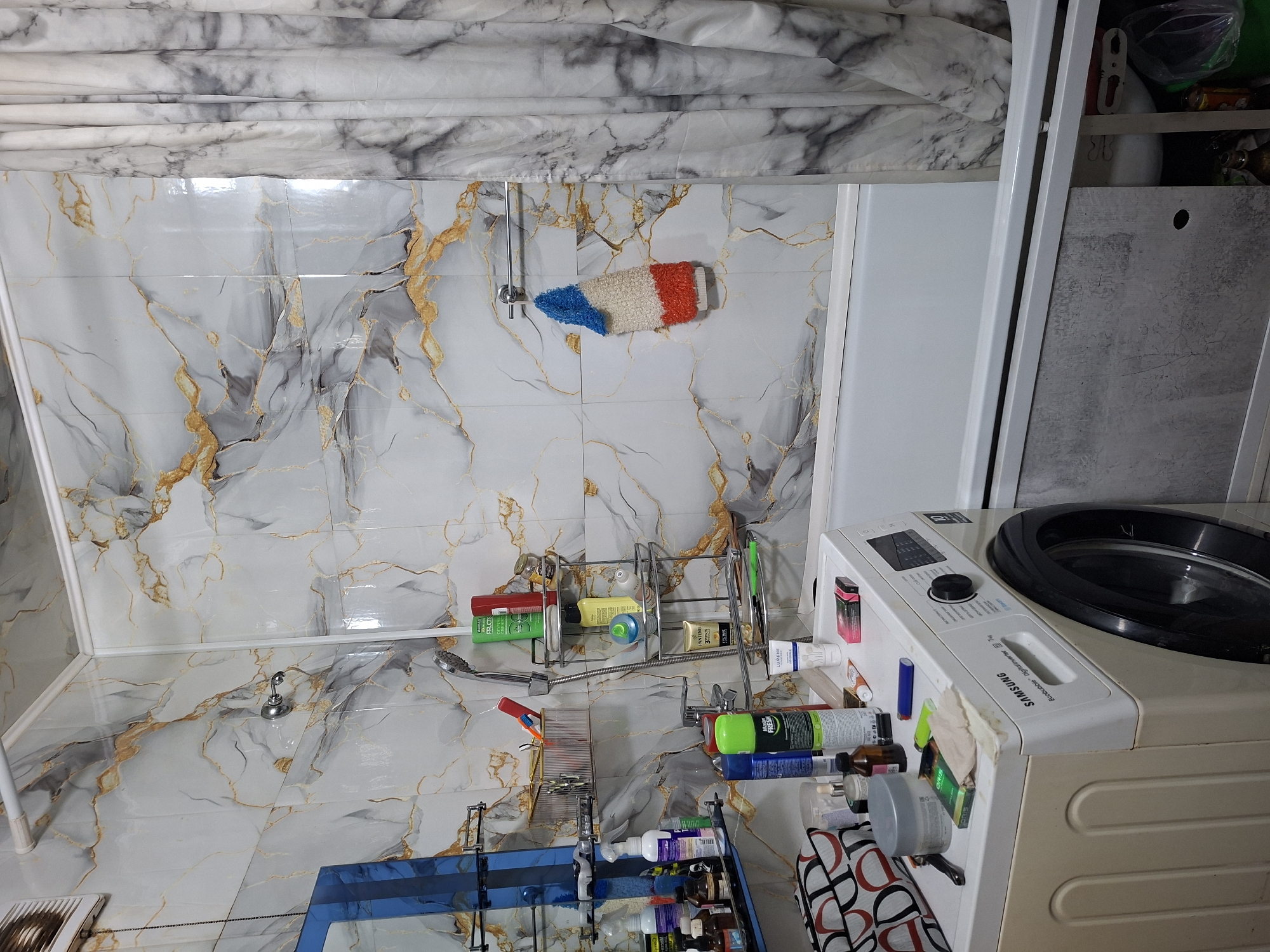 This is a medium shot of a cluttered bathroom in Ridder, Kazakhstan. The walls are tiled with a marble-like pattern featuring gold veining. A white bathtub is partially visible, with a grey and white marble-patterned shower curtain drawn to the right. To the left, a washing machine is positioned next to the bathtub. On top of the washing machine, there are various toiletries and cleaning supplies, including spray bottles, containers, and what appears to be a pack of cigarettes. A shelf unit is mounted on the tiled wall above the washing machine, holding more bottles and containers. A showerhead and faucet are visible near the shelf unit. A towel with red, white, and blue stripes hangs from a towel rack on the wall. The overall impression is of a functional but somewhat disorganized bathroom space.