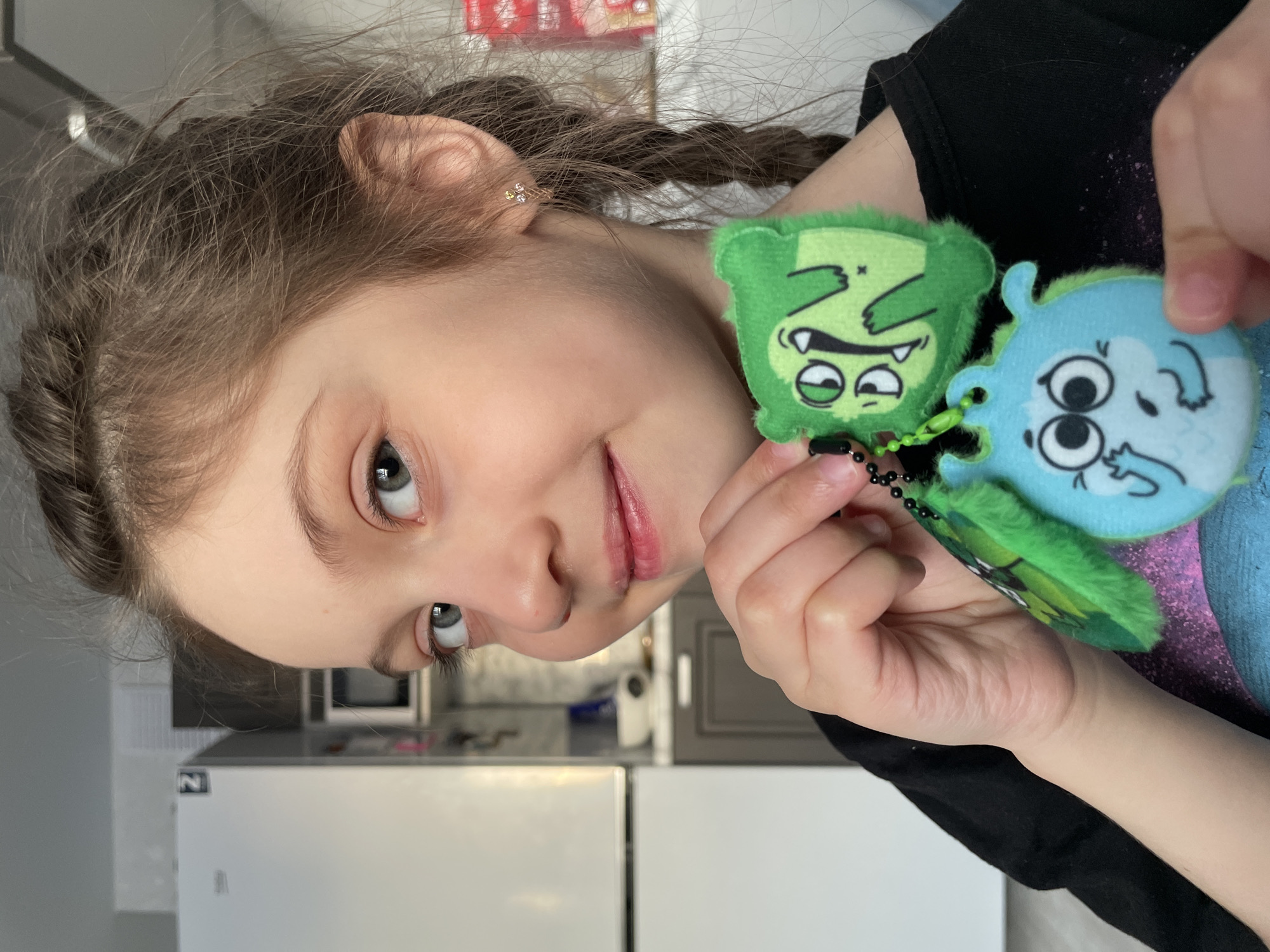A girl with braided brown hair and fair skin is holding two plush keychains in her right hand. The keychain on the left is green with a cartoon face, and the one on the right is blue, also with a cartoon face. The girl is wearing a black shirt. In the background, there is a kitchen with white cabinets, a microwave, and a box visible on a shelf. Based on the available information, it is not possible to determine if the scene is in Izhevsk, Russia.