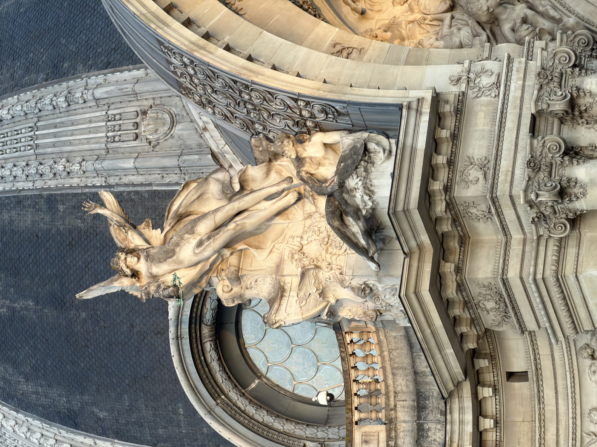 The image captures an architectural detail of a building in Paris, France. The scene is composed of intricately sculpted stone elements, including a trio of sculptures featuring human figures. The stone is a light cream color, showing subtle variations in tone and texture.
The sculptures appear to be allegorical, potentially representing figures from mythology or history. One sculpture, positioned prominently in the lower left corner, has wings and is partially draped. It leans outward, with one arm raised. Another sculpted group is visible to the right, depicting two figures in a classical style.
The surrounding architectural details are equally ornate, with decorative carvings and moldings. A large, rounded window is also visible, its glass panes reflecting the surrounding environment. The architectural style suggests a late 19th or early 20th-century construction. The background is a slate-covered roof.
A security camera is attached above the window. A small figure is barely discernible through the glass of the rounded window.