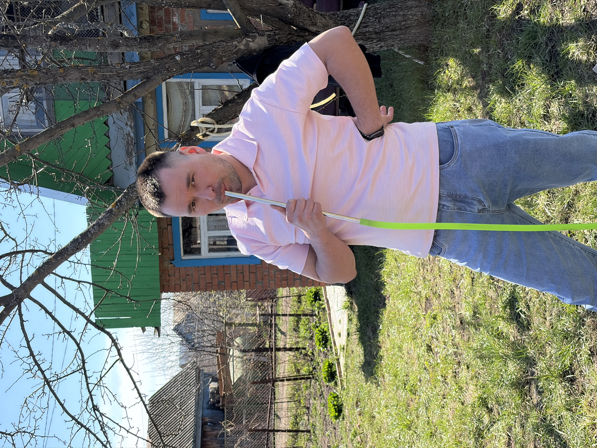 A man stands outdoors in a grassy yard, holding a long, green, flexible pipe to his mouth as if he is drinking or smoking through it. He wears a pink collared short-sleeved shirt and blue jeans. A residential house with a green awning and brick walls is behind him to the left, partially obscured by a bare-branched tree. In the distance, a fence and other buildings suggest a residential area. The location is likely in a rural or suburban part of Neftekamsk, Russia, indicated by the typical architecture and yard. The scene is brightly lit, suggesting a sunny day.
