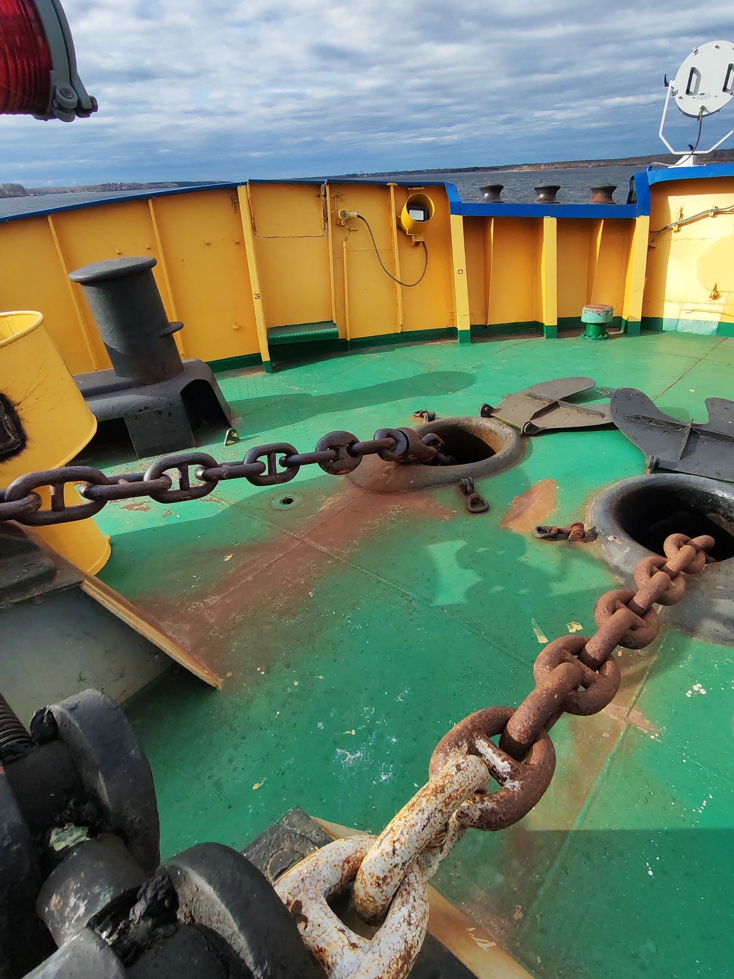 The image depicts the deck of a vessel, possibly a tugboat, at an undisclosed location. The deck is painted green and exhibits rust stains. Two open anchor chain wells are visible, with sections of heavy chain extending from them. The ship's superstructure is painted yellow with a blue trim. The sky is cloudy, and a distant shoreline is visible. The image does not show any people or specific actions. The location in Rybinsk, Russia, is not confirmed by the image.